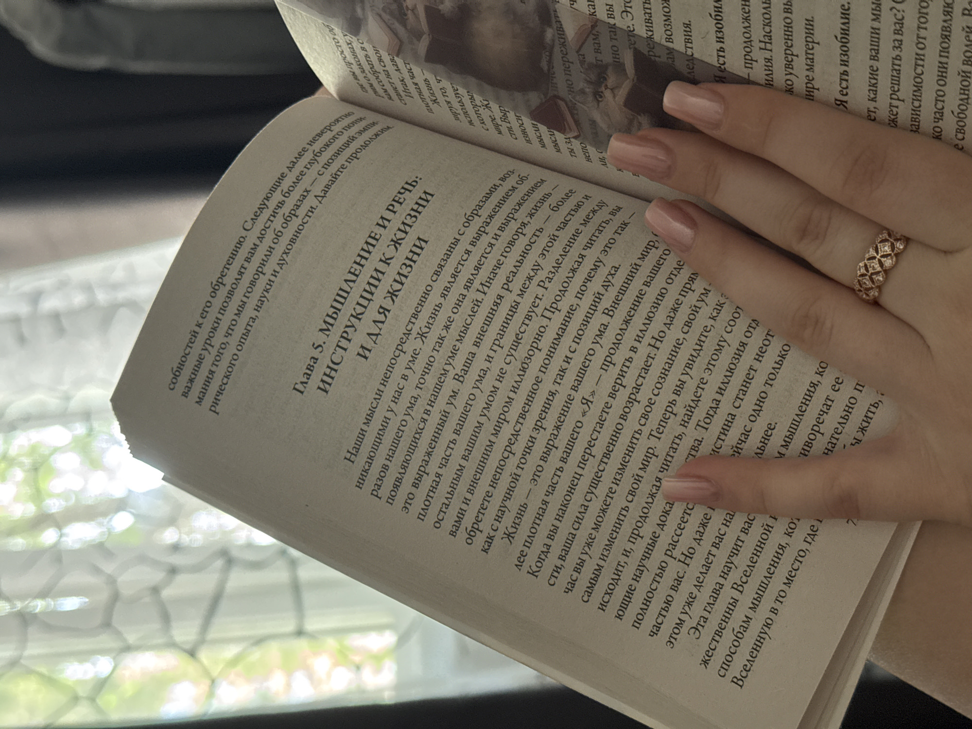 The image shows a close-up of a person's hand with manicured nails holding open a book. The book appears to be a paperback with text in Russian. The hand is positioned on the right side of the open book, with the fingers and thumb gently pressing down on the pages. A ring with a geometric design and small stones is visible on one of the fingers. The background is blurred and suggests an indoor setting with natural light coming from a window, possibly with greenery outside.
There is no information in the image that indicates the location is Novosibirsk, Russia.