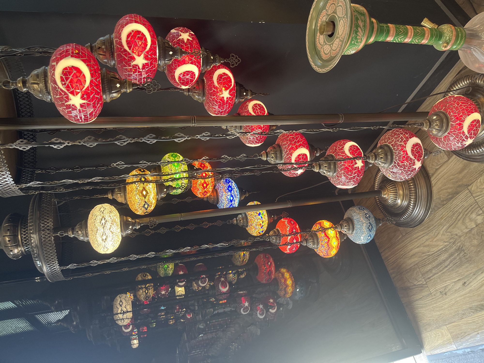 The image depicts a collection of ornate, mosaic glass lamps hanging from dark metal stands, alongside a decorated hookah. The lamps are of various colors, including red, yellow, blue, and white, and many feature a crescent moon and star motif, suggesting a Turkish influence. The background is dark, with a black wall and a wooden floor. The lighting is warm, emanating from the lamps themselves, creating a cozy and atmospheric effect. The scene appears to be an indoor setting, possibly a shop or a decorative display. The image does not provide any specific visual cues to suggest a location in Krasnodar, Russia.