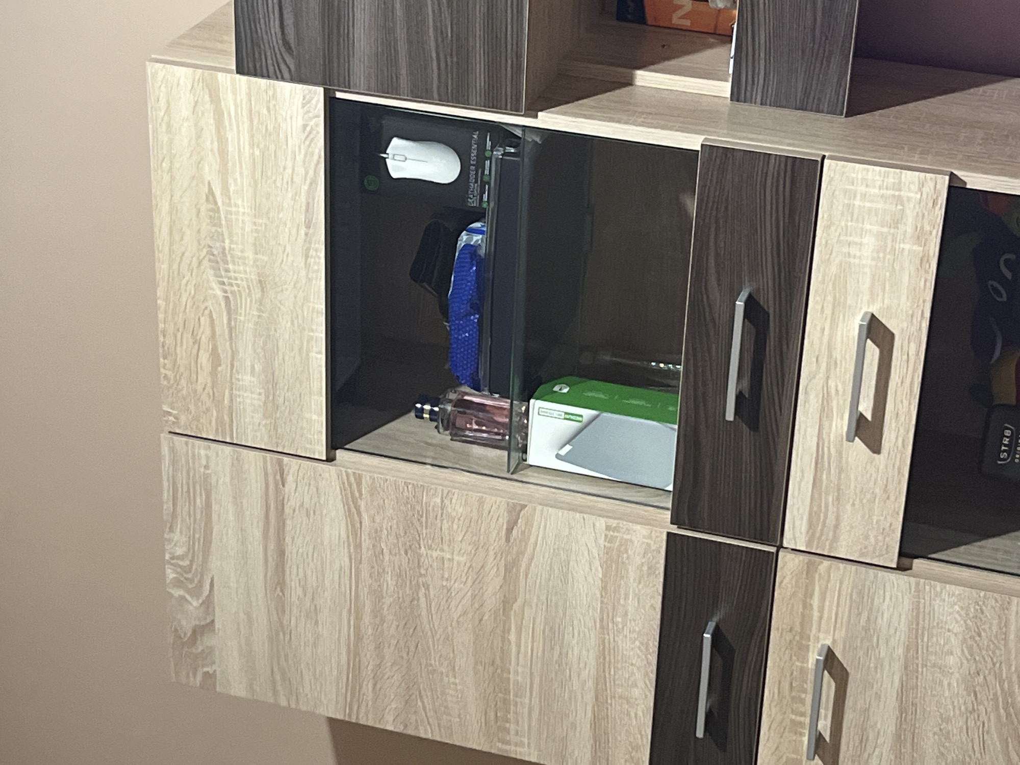 The image depicts a close-up view of a modern wooden furniture unit with multiple drawers and open shelving, indoors. The unit features a light wood grain finish combined with darker wood grain accents. In one of the open shelves, a pink bottle of perfume, a blue object, a white computer mouse in its packaging, a silver tablet, and a green box are visible. The drawers have silver handles. The lighting suggests an indoor environment, likely a domestic setting. The image does not contain any information that would allow for determination of location within the city of Dobrich, Bulgaria or any other specific geographic area.