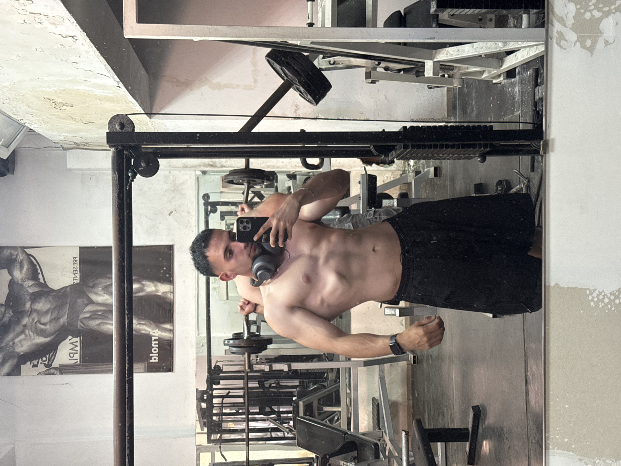 A young man, with dark hair and stubble, stands in a dimly lit gym, taking a mirror selfie. He is shirtless, revealing a muscular physique, and wears black athletic shorts and a black wristwatch on his left wrist. Headphones rest around his neck. The gym appears old and worn, with peeling paint on the walls and exposed concrete. Weights and gym equipment are visible in the background, including a barbell with weights, weight machines, and benches. A poster of a muscular man is displayed on the wall. The floor is concrete and appears dusty. The overall impression is of a functional but unkempt workout space. The location is specified as a gym in the city of Shumen, Bulgaria.