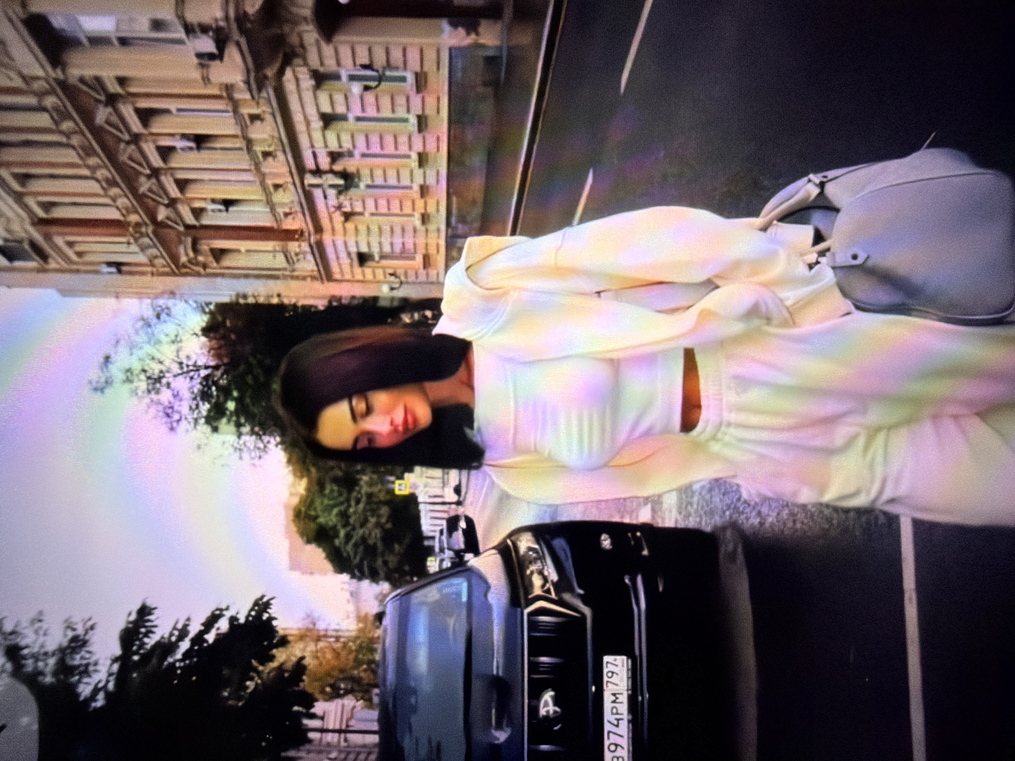 In a street scene in Beșalma, Moldova, a young woman with dark hair and closed eyes is depicted. She is wearing a light-colored, pastel tie-dye loungewear set, consisting of a cropped hoodie and matching pants. A light grey handbag rests on her shoulder. She appears to be leaning against a black vehicle, which is parked on the street. The vehicle's license plate is visible, displaying the text "3974PM 797".
Behind the woman and the car, an old, multi-story building with numerous windows is visible, suggesting an urban or semi-urban environment. Trees with green and autumnal foliage are also present, some in the foreground and others behind the building. A faint, pastel-colored rainbow or light effect is visible in the sky, adding a touch of color to the scene. The image appears to be taken during daylight, possibly late afternoon or evening, given the lighting conditions and the presence of the rainbow effect. The woman's closed eyes and relaxed posture suggest a moment of quiet or repose.