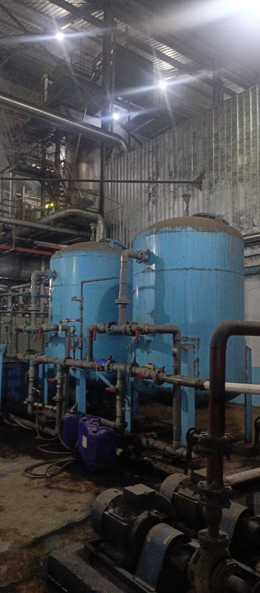 This is a medium-shot, vertically oriented, indoor photograph taken at the Iemirah, Indonesia water treatment facility. The scene depicts industrial machinery, specifically two large, blue cylindrical tanks that appear to be water filters or pressure vessels. These tanks are connected by a complex network of pipes, valves, and smaller tanks, indicating an active operational system. The machinery is positioned on a dirty concrete floor, with visible electrical conduits and motors in the foreground, suggesting a functional industrial setting. Overhead, a metal roof structure is visible, supported by beams and industrial lighting fixtures that cast strong beams of light, creating lens flare effects. The overall environment appears somewhat aged and industrial, with dust and wear visible on the surfaces. There are no people visible in the photograph.