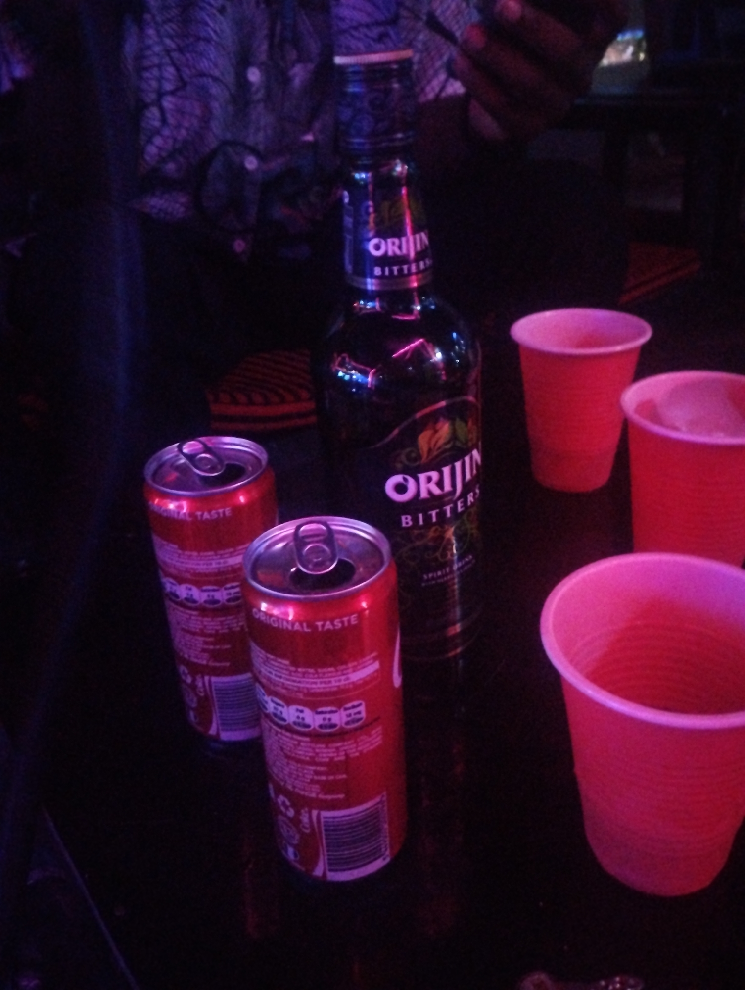 The image shows a table with two open cans of Coca-Cola, a bottle of Orijin Bitters, and three red plastic cups. The location is likely a bar or social gathering, possibly in Tumfure, Nigeria, suggested by the Orijin Bitters bottle (a popular Nigerian beverage). A person's hand holding a phone is visible in the background. The environment is dimly lit, with purple lighting.