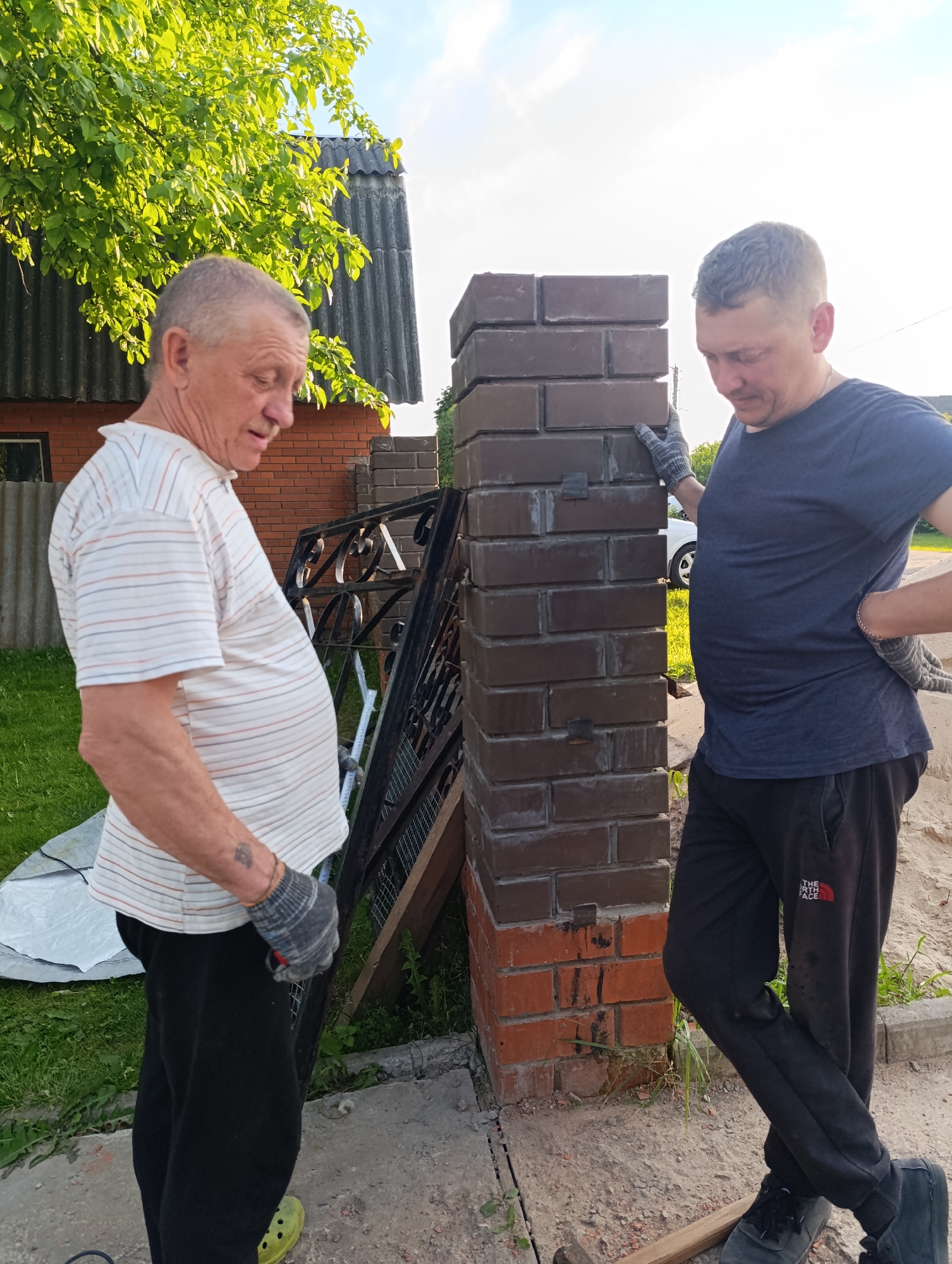 The image depicts two men engaged in an outdoor construction or installation project. The man on the left is older, with short grey hair and a mustache, wearing a white striped polo shirt, black pants, and green clogs, along with grey work gloves. A tattoo is visible on his right forearm. The younger man on the right has short dark hair and is dressed in a dark blue t-shirt, black sweatpants, and dark shoes, also wearing grey work gloves.
They are standing next to a brick pillar, which is constructed from brown bricks at the upper levels and reddish-orange bricks at the base. A black, ornate metal gate or fence section is propped leaning against the pillar and a wooden frame in the background. In the deeper background, there is a building with a red brick wall and a grey corrugated roof, partially obscured by a vibrant green tree. On the ground, a pile of sand and a grey tarp are visible, indicative of a work site. A white car is partially visible behind the younger man.
Both men are looking downwards, appearing focused on the task at hand. The bright natural lighting and the presence of shadows suggest it is either late afternoon or early morning. The setting appears to be a residential or rural outdoor area.
The younger man's black sweatpants feature the "THE NORTH FACE" logo in red and white on the left thigh.