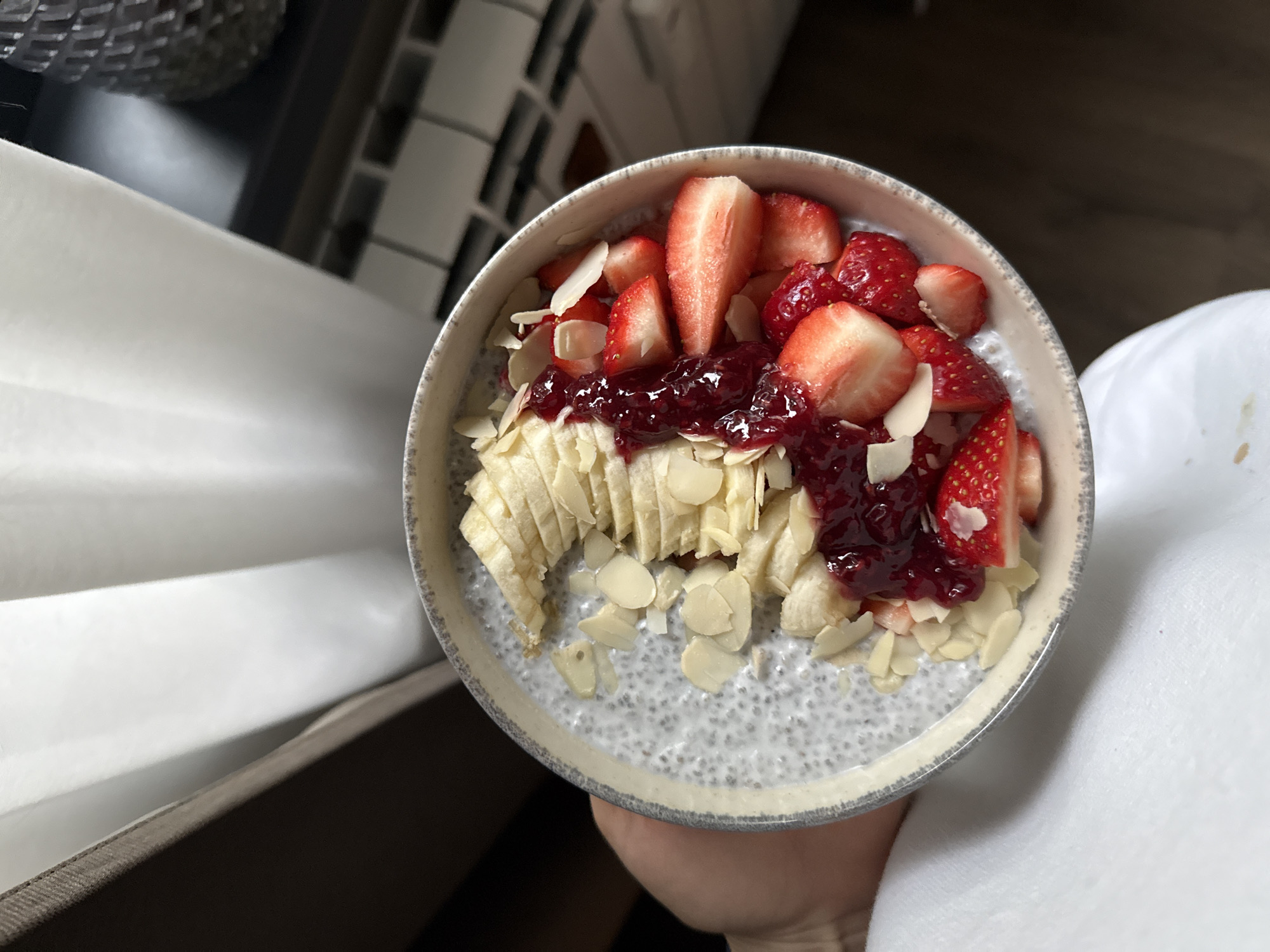 The image shows a close-up, high-angle view of a bowl filled with a breakfast dish, possibly chia seed pudding, topped with sliced strawberries, banana slices, jam, and almond flakes. The bowl is held by a person wearing a white top. The background includes a blurred area, possibly a counter or table near a window. It is impossible to determine if this image was taken in Sofia, Bulgaria, as there are no elements in the image that would identify the location.