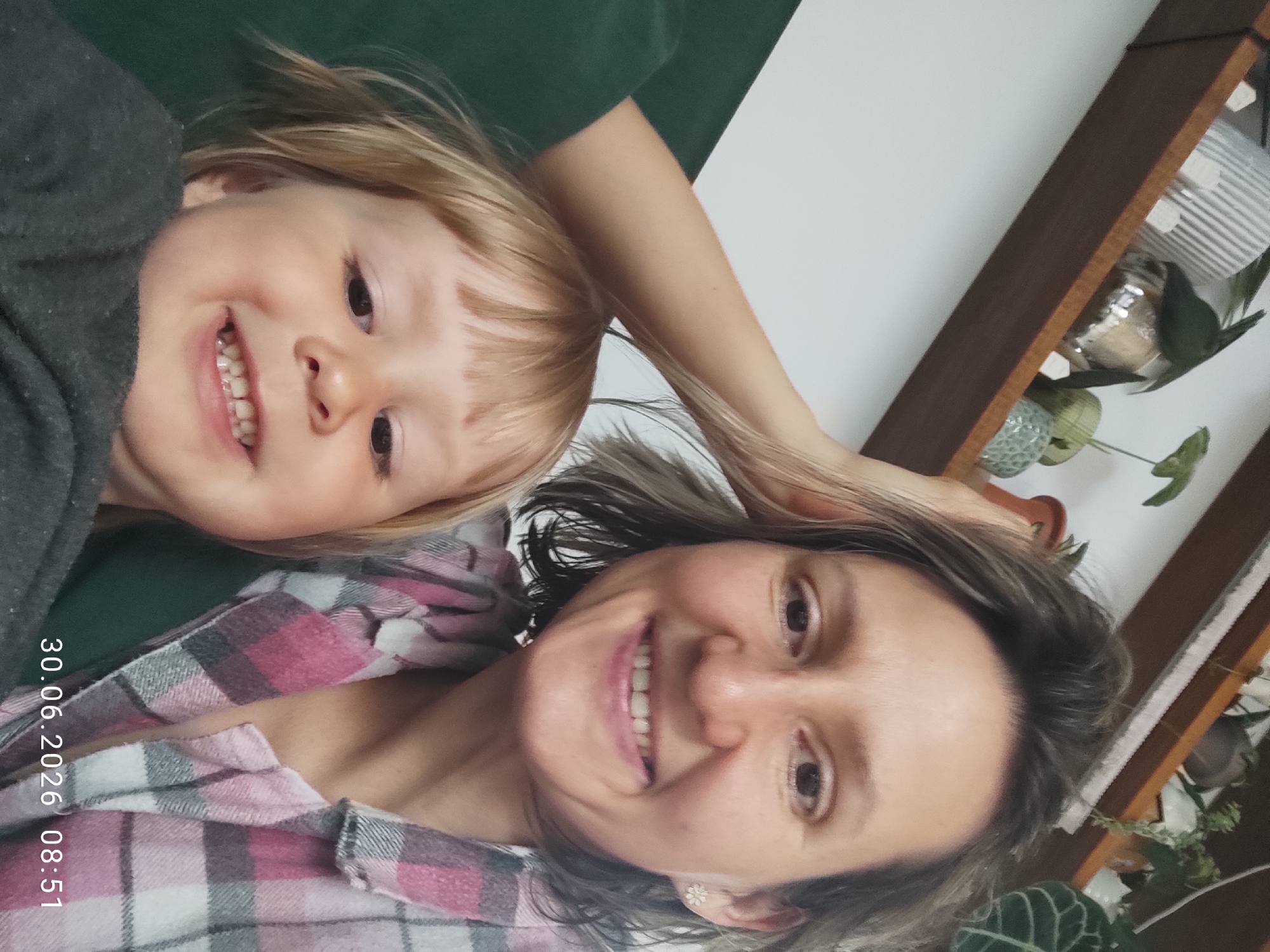 In the foreground of the image, a woman and a young child are taking a selfie. The woman, on the right, has brown hair and is smiling at the camera. She is wearing a pink and grey plaid shirt. The child, on the left, has blonde hair and is also smiling. The background shows a wooden shelf filled with plants in pots. The overall environment appears to be indoors. The timestamp "30.06.2026 08:51" is visible in the lower right corner of the image. There is no information to suggest this image is related to Malyi Bukor, Russia.