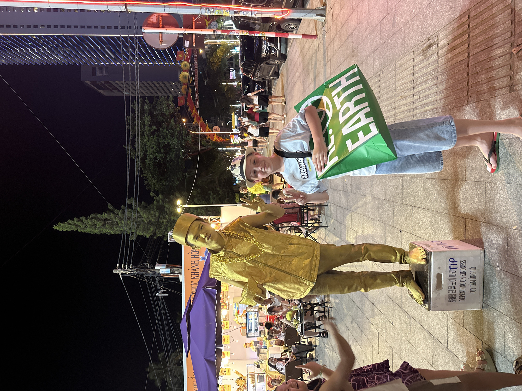 The image depicts a lively night scene on a street in Nha Trang Ward, Vietnam, suggesting a bustling night market or entertainment area. In the foreground, a street performer, entirely painted in gold to resemble a living statue, stands on a white box. He wears a golden hat, vest, pants, and a thick gold chain, holding a golden object while striking a pose with a raised hand. The box serves as a tip collection point, displaying text in Korean ("친절도에 따라 TiP"), English ("DEPENDING ON KINDNESS"), and Vietnamese ("TÙY TÂM ỦNG HỘ"), along with a QR code.
Beside the performer, a young boy in a patterned hat, light t-shirt, and jeans holds a large green bag with the text "EARTH" and "ORDINARY IS NOT FOR US." He looks towards the camera with a smile. In the background, numerous people are visible, some seated at tables under purple awnings, indicating food stalls or outdoor dining. A banner in the mid-ground reads "THỰC KHÁNH HÒ," suggesting a food zone within the Khanh Hoa province.
The scene is well-lit by streetlights and the glow from various establishments against the dark night sky. Vietnamese flags and other festive decorations are visible along the street. Overhead utility wires and distant buildings add to the urban setting. The atmosphere appears vibrant and engaging, characteristic of an evening street gathering.