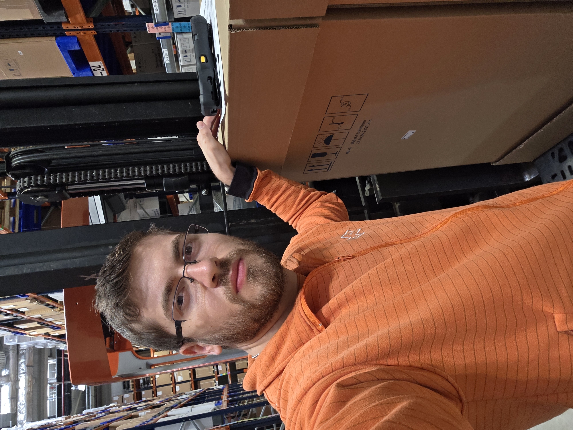 A man is standing in a warehouse, likely in Plovdiv, Bulgaria, though this cannot be confirmed with visual data. He is holding a handheld barcode scanner to a cardboard box on a forklift. The man is wearing glasses and an orange zippered sweatshirt. Shelves of inventory are visible in the background.