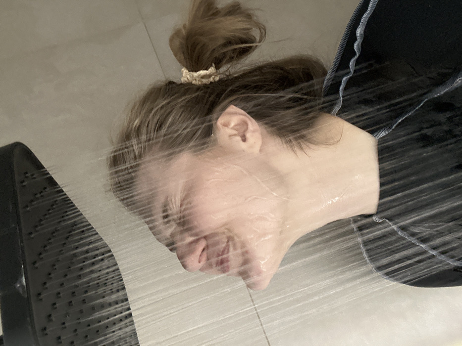 A person's head and upper body are depicted under a stream of water. The individual has fair skin and light brown hair styled in a bun, held with a scrunchie. The person wears a dark-colored top. Water droplets and streaks cover the face, distorting the features. To the left, a showerhead is visible, indicating the source of the water. The background appears to be a tiled bathroom or shower area, with light-colored tiles partially visible.