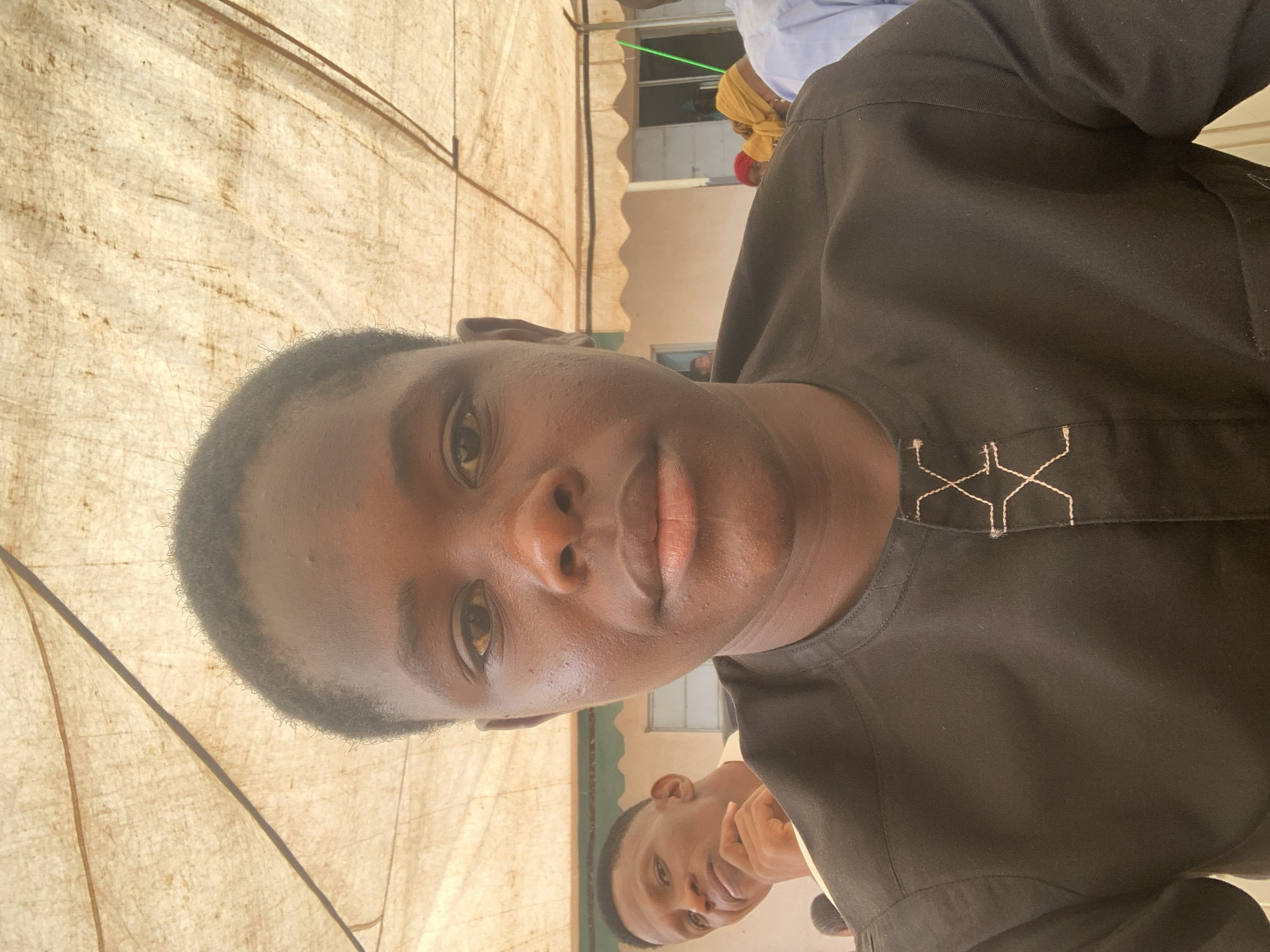 A close-up selfie shows a young man with dark brown skin and short dark hair. He is wearing a dark, possibly black, collared shirt with light-colored geometric embroidery on the collar. A second person, another young man with similar features, appears blurred in the background towards the bottom. The selfie is taken outdoors under a fabric covering, possibly a tent or awning, with visible supports. Other people are visible in the background beyond the fabric covering. The image location is suggested to be in Jos, Nigeria, although no specific landmarks are visible.