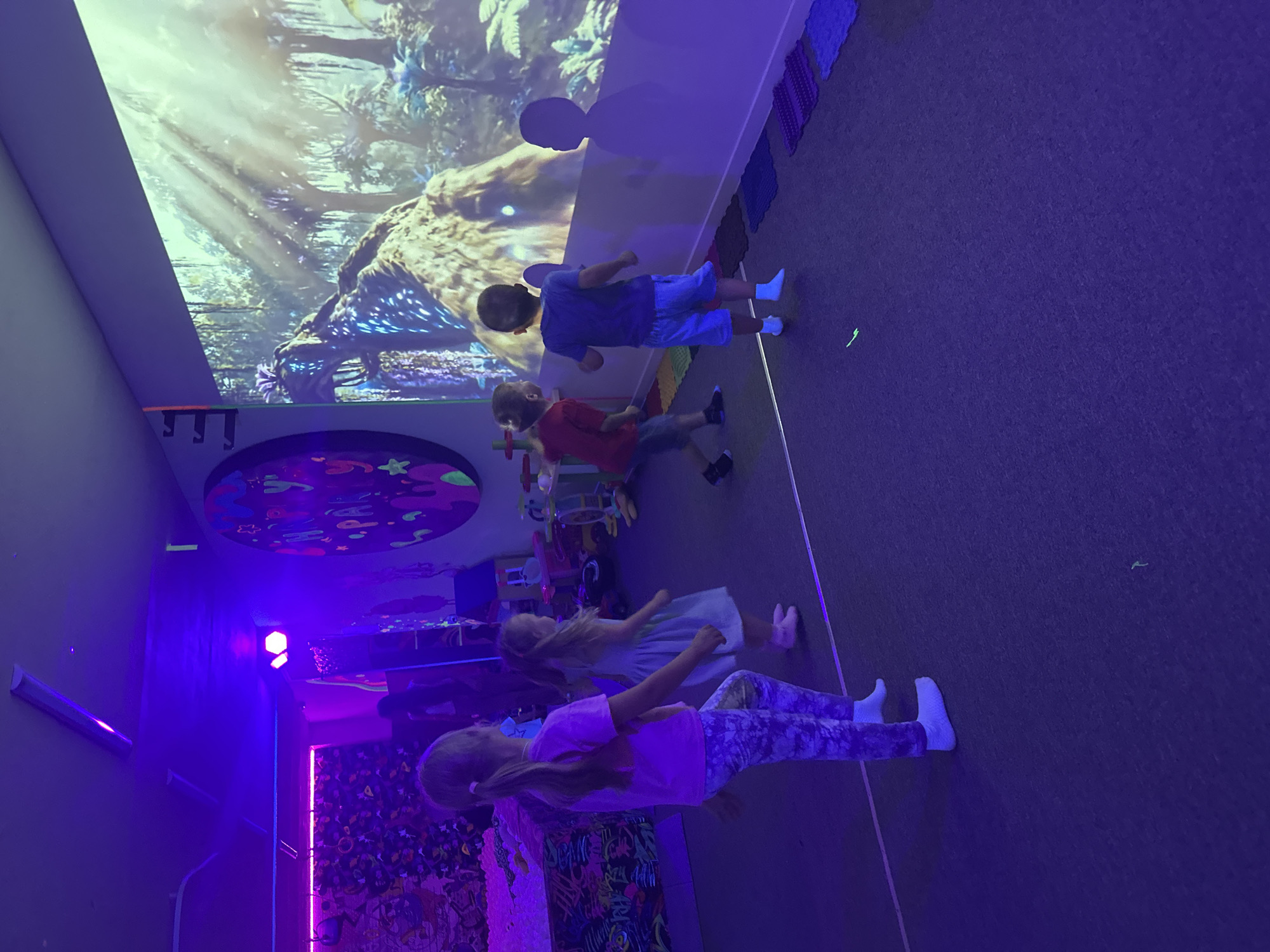In an indoor entertainment space illuminated by purple and blue lights, four children stand facing a large projected image of a jungle scene with a dinosaur-like creature. Two girls, one in a pink shirt and patterned leggings, and another in a light blue dress, are positioned on the left. Two boys, one in a red shirt and grey shorts, and another in a blue t-shirt and blue shorts, are on the right. The floor is a dark carpet with a line dividing it. Several colorful mats are arranged near the wall on the right. The room appears to be a children's play area, possibly a party venue, with toys visible in the background.