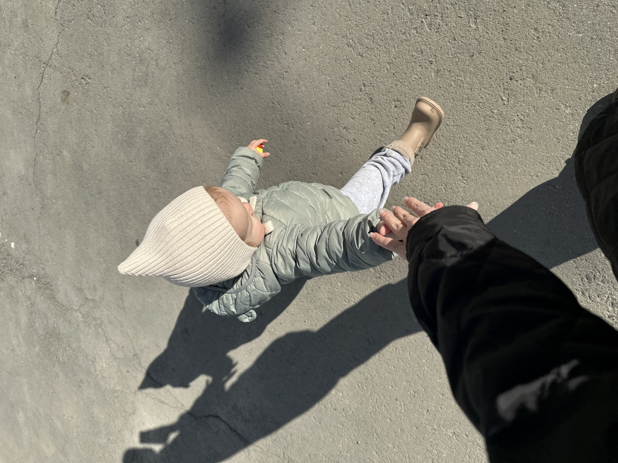 A top-down shot shows a child in a light green puffer jacket, gray sweatpants, beige hat, and beige boots. The child holds a small yellow object in their left hand. An adult's hand reaches out towards the child. The scene appears to be outdoors on a concrete surface. No location is discernible from this photo.