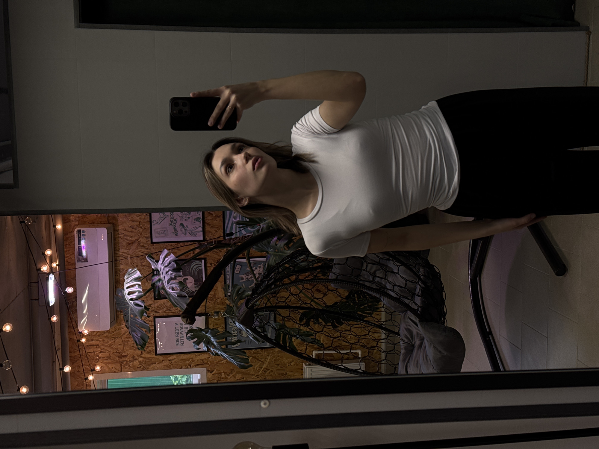 A young woman is captured in what appears to be a mirror selfie, standing in an indoor setting. She has shoulder-length brown hair and is wearing a white short-sleeved t-shirt and black pants. Her right arm is raised, holding a dark smartphone with a visible multi-lens camera, pointed towards herself. Her gaze is directed slightly upwards and to her left, not directly at the phone's lens.
The background behind her features a rustic wooden wall adorned with string lights containing numerous small, round bulbs. An air conditioning unit is mounted on this wall, with some light reflected on its surface. Below it, a narrow horizontal window is visible. Several framed posters or artworks decorate the wooden wall; one poster, partially obscured, appears to have the inverted text "CLINIC". Large, artificial Monstera leaves are interspersed among the decorations. To her right, part of a dark, net-like hanging chair with a metal frame is visible. The floor is light-colored, possibly tiled, and a dark table or surface is partially seen in the lower right corner, indicating she is near furniture.
The lighting is dim and warm, contributing to a cozy or relaxed atmosphere. The image is taken indoors, likely in an establishment such as a cafe, lounge, or a decorated room, indicated by the specific decor elements. No specific time of day is discernible, but the indoor lighting suggests evening or an intentionally controlled ambiance.