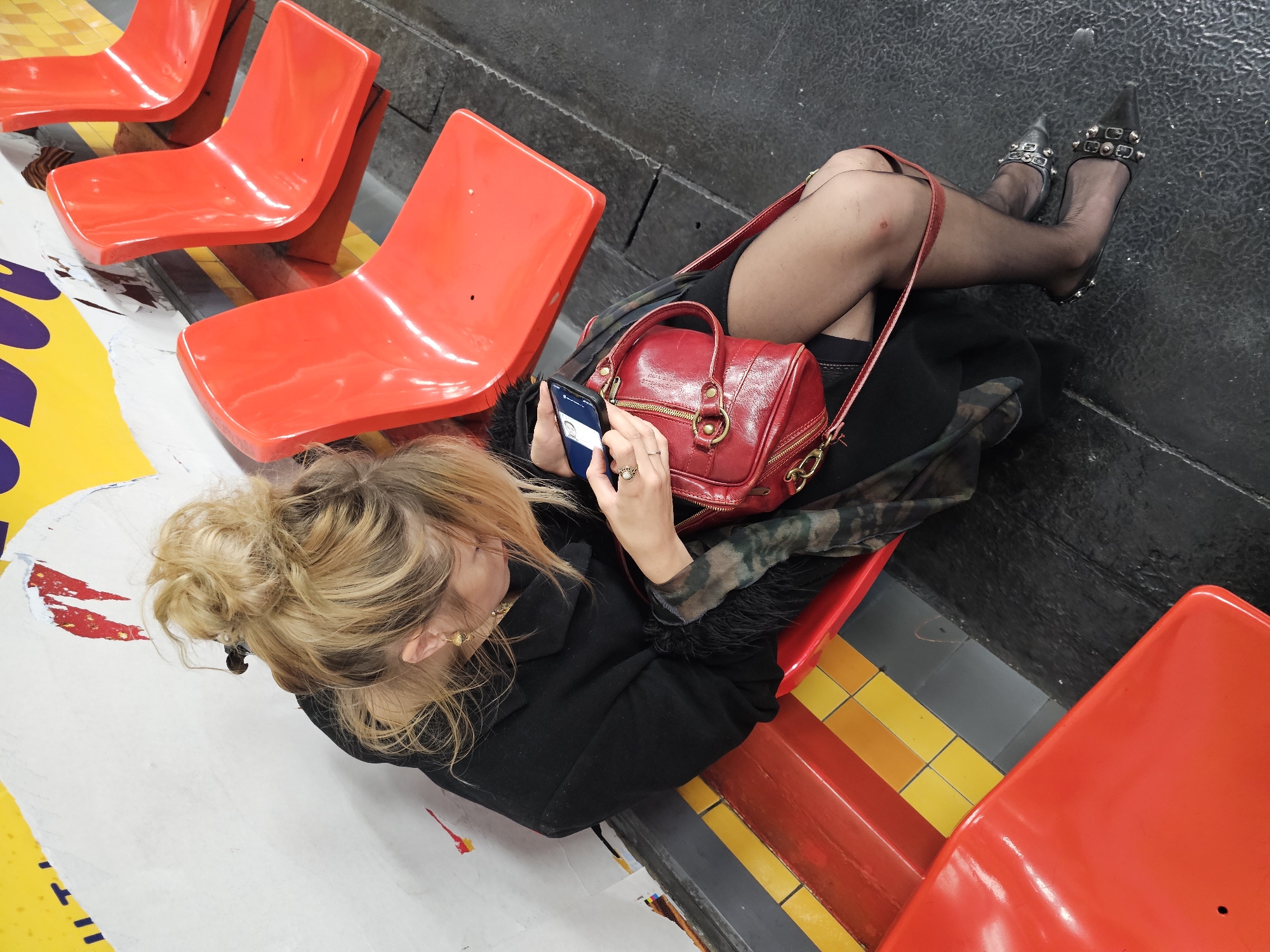 A high-angle shot depicts a woman seated in a public area. She has blonde hair styled in a bun and is wearing a black coat. She is looking at her smartphone, which she holds in her hands. A red leather bag sits on the seat beside her. She is wearing black tights and embellished high heels. The seating consists of bright red plastic chairs. The flooring is made of yellow and gray tiles, with painted artwork on parts of the floor. The scene is set on a subway platform in the city of Paris, France.