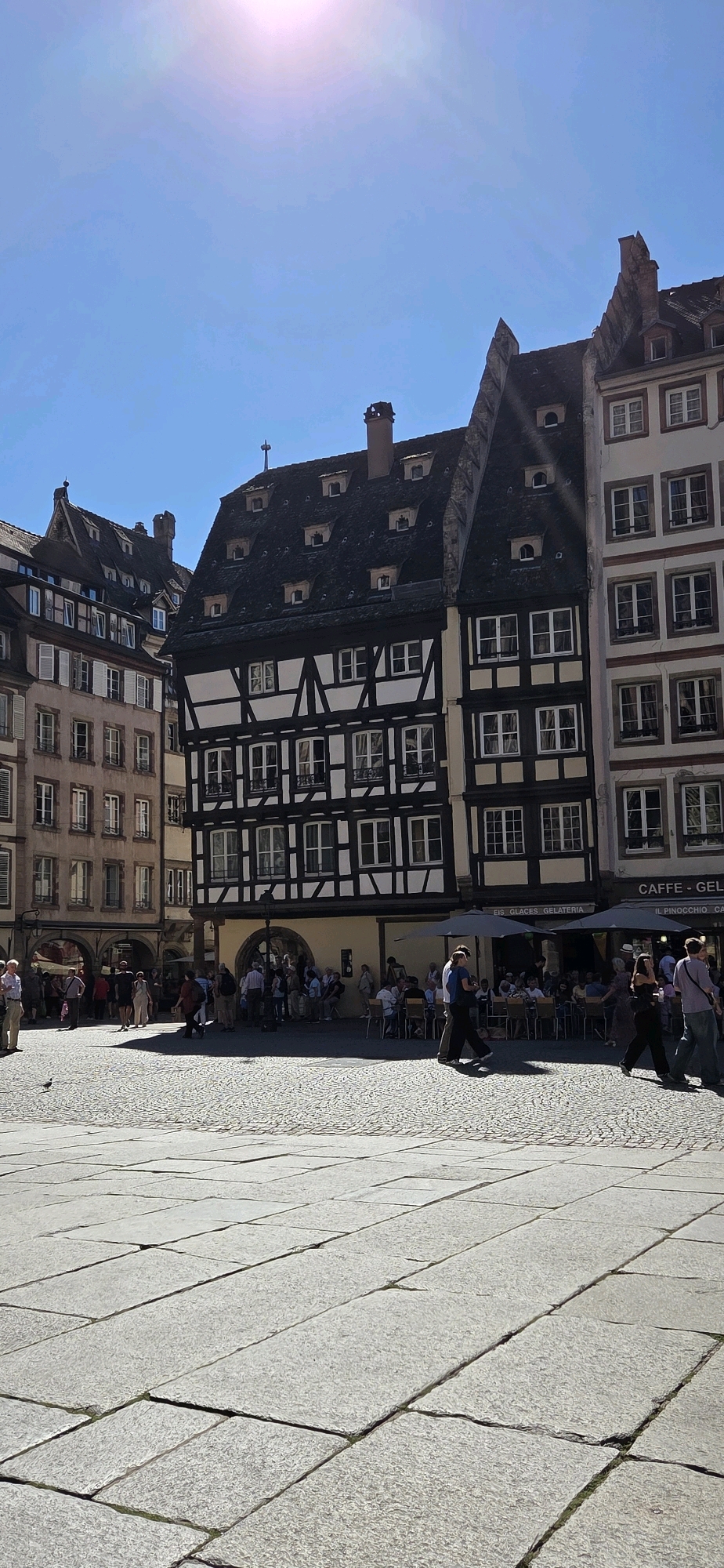 The image captures a sunny day in a European city square, likely in Strasbourg, France, given the distinctive half-timbered architecture. In the center, a prominent building features a dark, steep roof with numerous dormer windows and a classic half-timbered facade of black beams against a cream-colored background. To its left, a taller building with a lighter facade and shuttered windows stands, while to its right, another building with a similar facade and multiple windows is visible. The square itself is paved with large, irregular flagstones in the foreground and smaller cobblestones further back. Several people are scattered across the square, some walking and others seated at outdoor tables belonging to a cafe visible beneath umbrellas. A small bird is perched on the paving stones in the lower left. The sky is a clear, bright blue, with the sun casting distinct shadows. The scene depicts a vibrant public space bustling with activity under a clear sky.