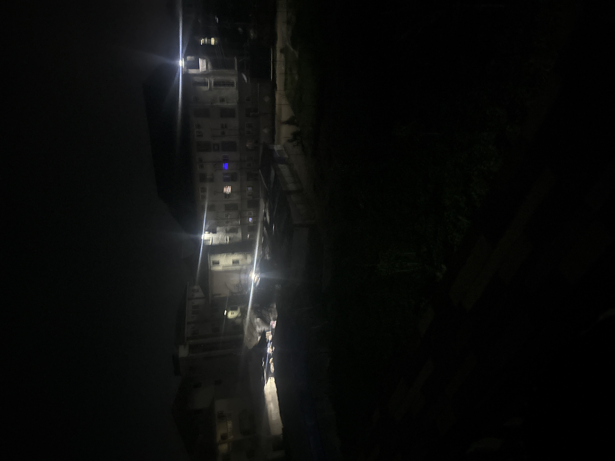 The image shows a building at night in Ihiagwa, Nigeria. The building is tall and appears to have multiple floors, with several windows visible, some illuminated. The area surrounding the building is dark, with some vegetation visible on the right side of the frame. The image is taken from a high vantage point.