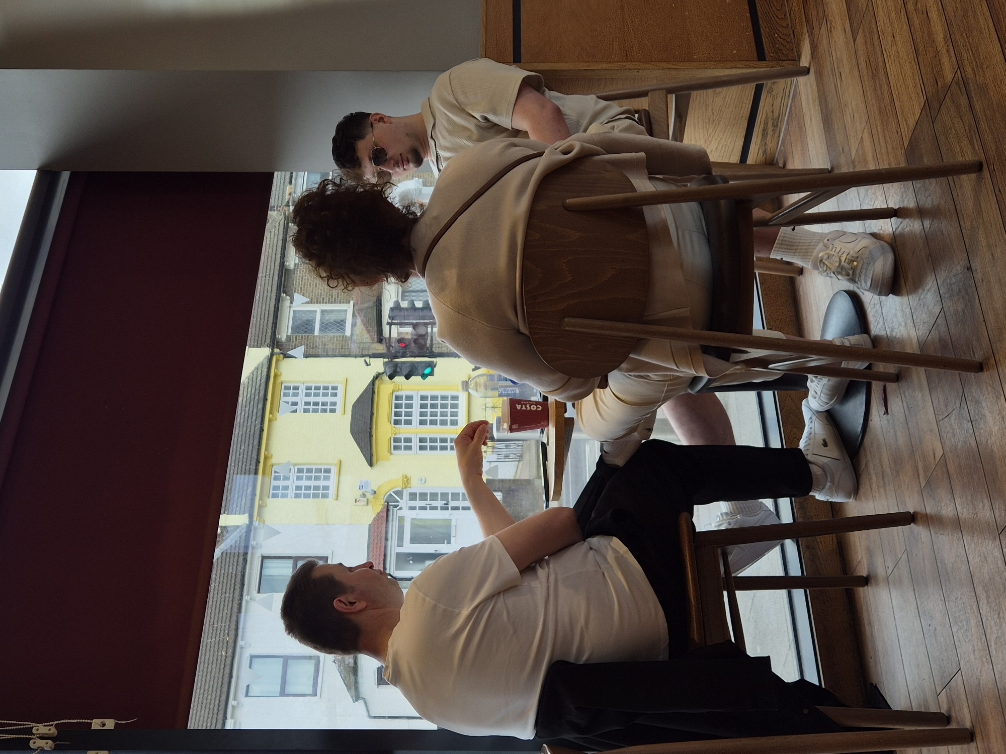 Three people are seated indoors, likely in a café, next to a large window. The person closest to the window is wearing a white t-shirt and black trousers. The middle person has curly hair and is wearing a beige jacket. The third person, partially visible, wears sunglasses and a beige top. A Costa coffee cup is visible on the table. Outside the window, buildings, including a yellow building, are visible; a traffic light is also seen. The location is potentially in London, United Kingdom, given the architectural style. The interior has wooden flooring.