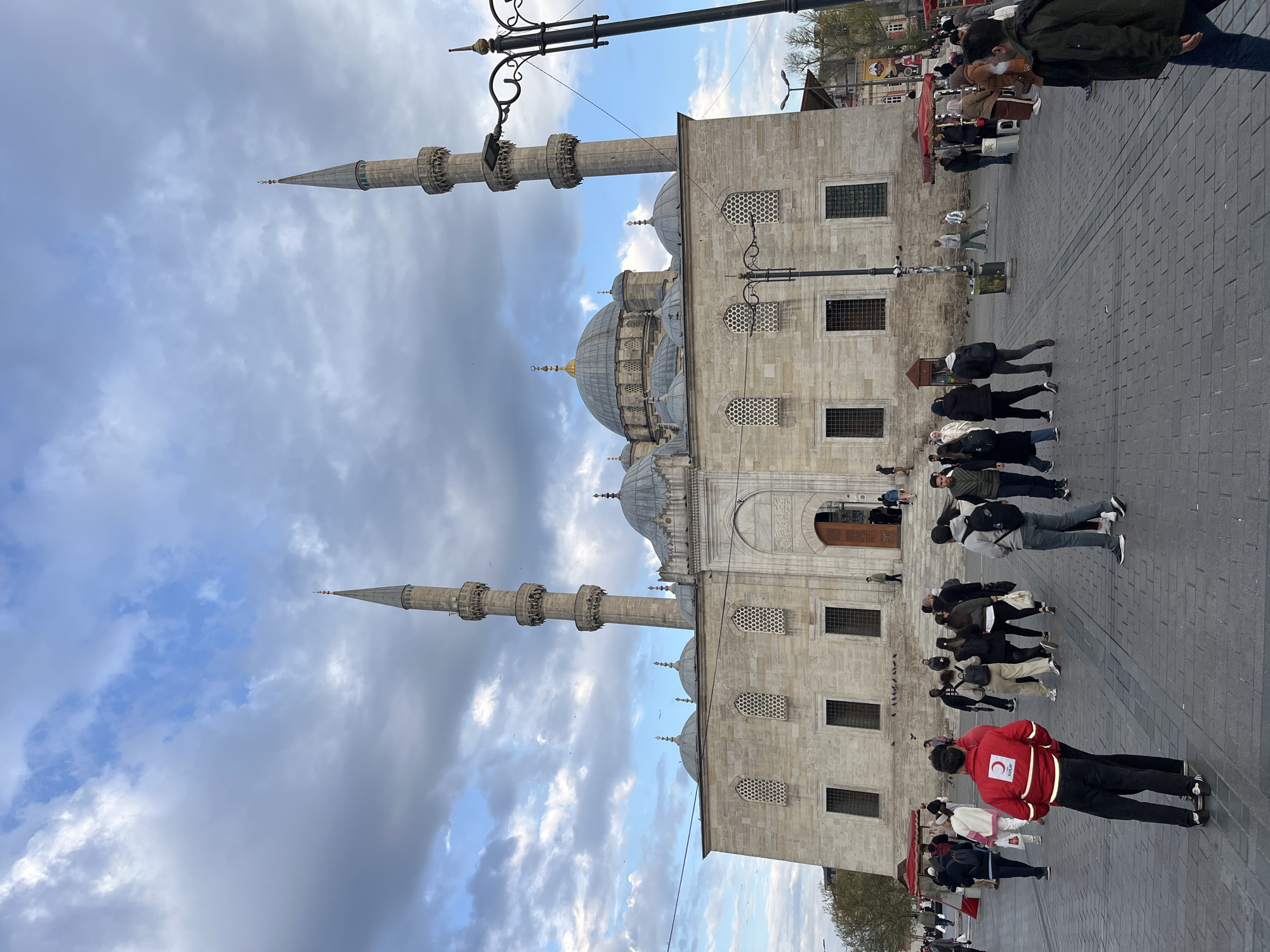 A wide shot captures a grand mosque and its surrounding plaza under a cloudy sky in Istanbul, Turkey. The mosque, constructed from light-colored stone, features a large central dome flanked by smaller domes and two tall, slender minarets with multiple balconies and conical spires. Its facade is adorned with decorative arched windows and rectangular window openings.
In the foreground and mid-ground, numerous people are scattered across a paved public square. Most individuals are seen walking or standing, dressed in casual attire. A prominent figure in the foreground wears a bright red jacket with a white crescent moon and star emblem (Kızılay logo) on the back, facing away from the camera. Other smaller figures can be seen near what appear to be market stalls or kiosks further in the background to the left of the mosque. A decorative black lamppost extends into the upper left of the frame.
The sky is overcast with a mix of grey clouds and some patches of blue, suggesting daytime. The overall impression is a busy urban scene centered around a significant architectural landmark.