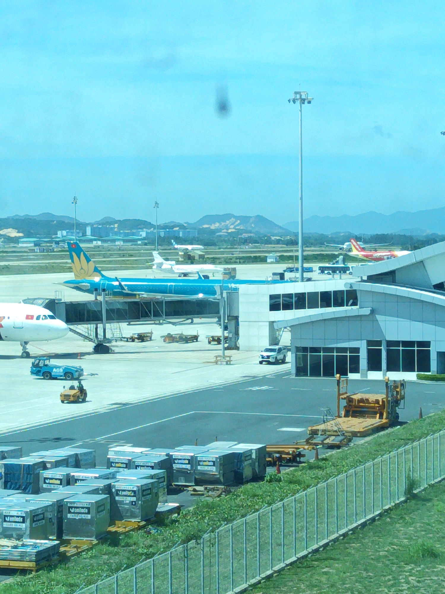 The image depicts an airport tarmac at Bắc Cam Ranh, Vietnam. Two airplanes are visible: one is a blue and gold Vietnam Airlines aircraft connected to a jet bridge, and another is a white aircraft parked to its left. In the background, a red and yellow airplane is visible near a terminal building. Various airport vehicles, including a blue baggage tug with "AGS" written on it and a yellow tow tractor, are present on the tarmac. Several cargo containers are stacked on pallets in the foreground, with a chain-link fence bordering a grassy area. The sky is bright blue with some scattered clouds, and mountains can be seen in the distance. The overall scene suggests a busy airport environment.