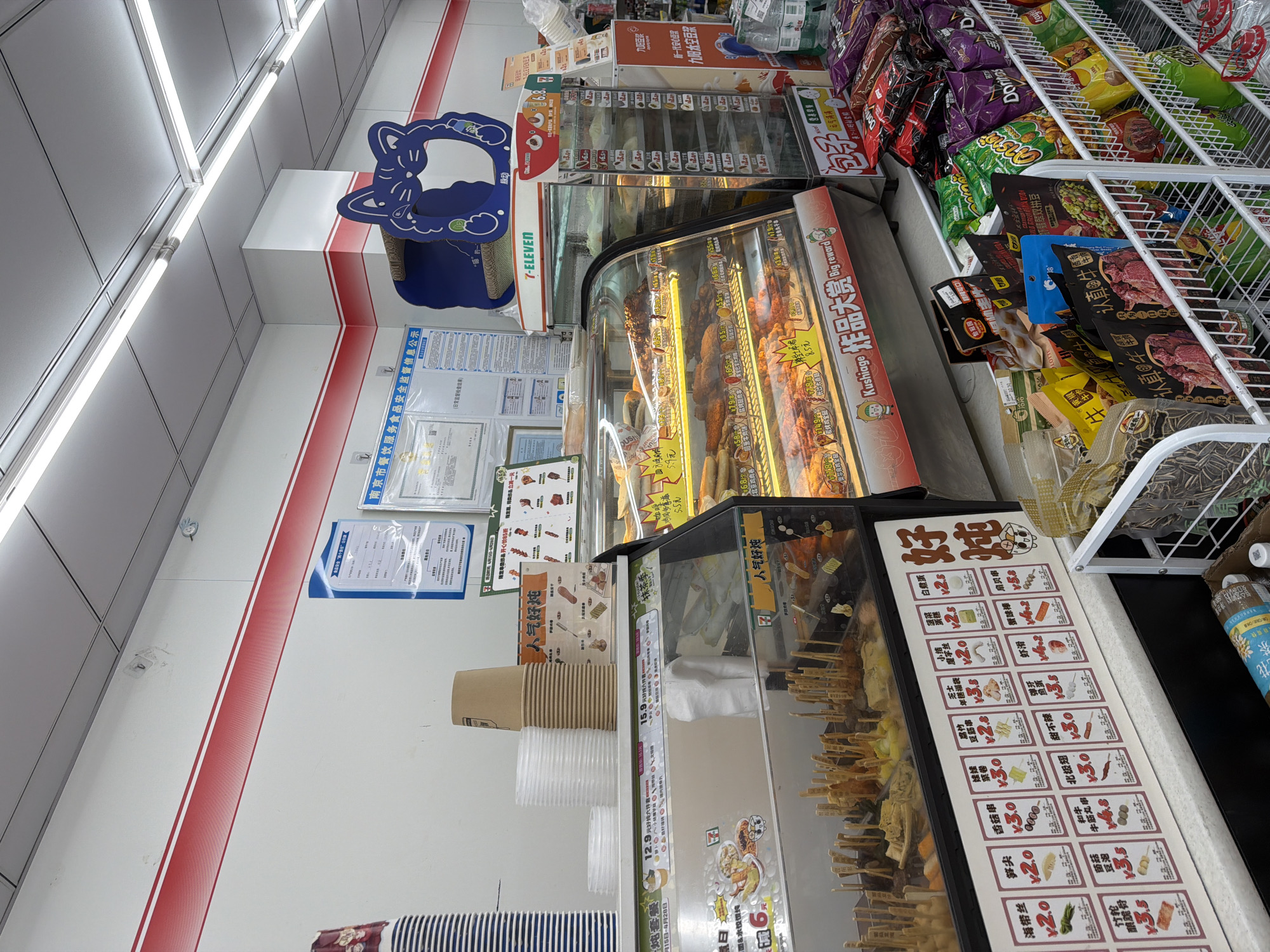 Here's a detailed description of the image:
**Location:** Jiangning District, Nanjing, China.
**Scene:** The interior of a 7-Eleven convenience store.
**Objects:**
* Refrigerated display cases filled with a variety of ready-to-eat foods.
* Snack items (chips, dried meat) on shelving.
* Hot food items, including skewers and fried foods, are visible in the display cases.
* Paper cups stacked near a display of "Kushiage" (fried skewers).
* Promotional signage for food items, including prices.
* Information posters about food safety regulations.
* 7-Eleven branded signage and logos.
* Packaging for various beverage brands like "Jiuyang Soymilk".
**Subjects:** The focus is on food items and store displays rather than people.
**Actions:** The image depicts food items presented for sale.
**Environment:** The store is brightly lit with fluorescent lights. The store is clean and well-organized. The color scheme of the store primarily consists of white, red, and black.