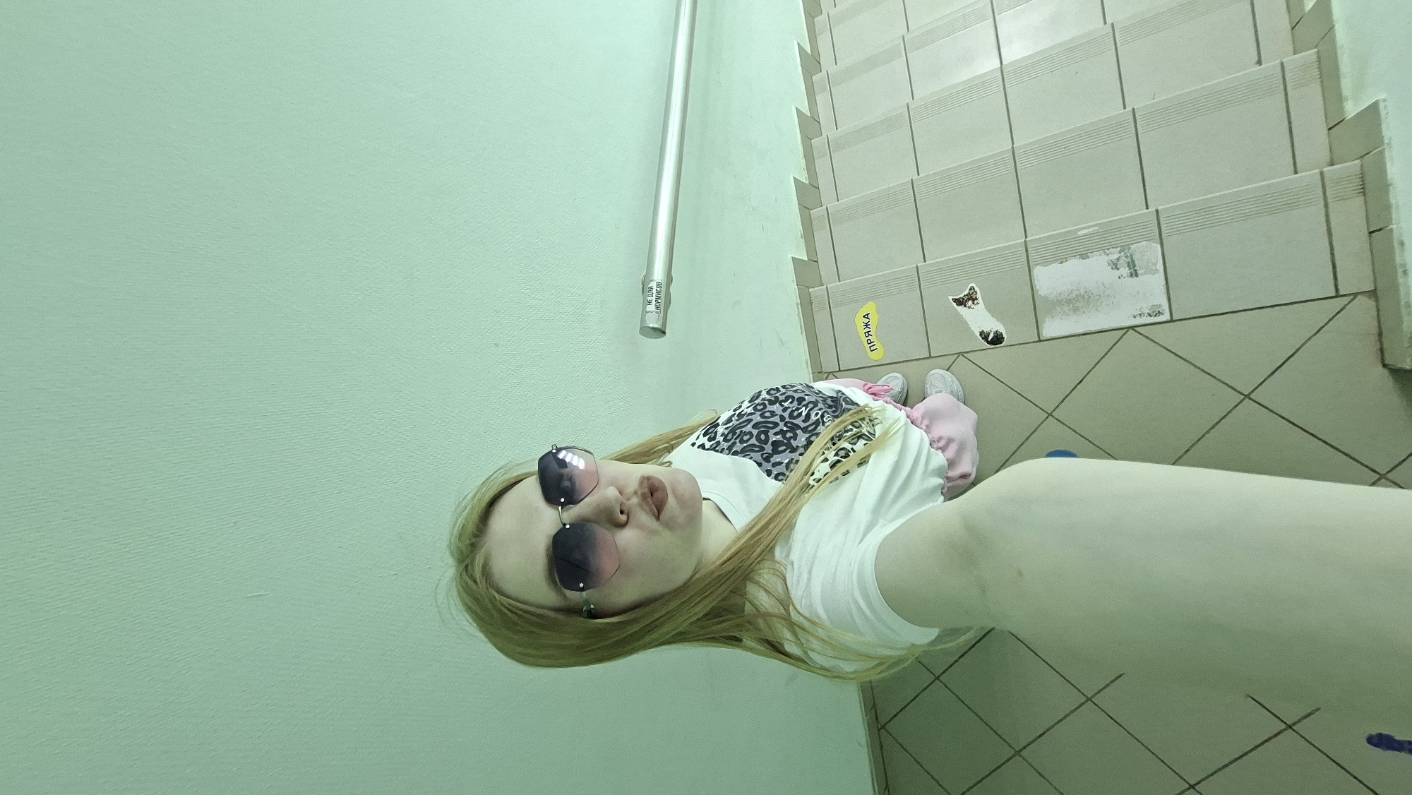 A woman with long blonde hair is taking a selfie in what appears to be an indoor stairway. She is wearing sunglasses, a white t-shirt with a leopard print design, and pink pants. Her right arm is outstretched towards the camera. The stairway is tiled with light-colored, rectangular tiles on the steps and square tiles on the landing. There is a metal handrail attached to a light green wall. A yellow sticker with Russian text is visible on one of the steps. A sticker in the shape of a sock is on the floor. Based on the writing near the handrail, the location is likely Chaikovsky, Russia.
