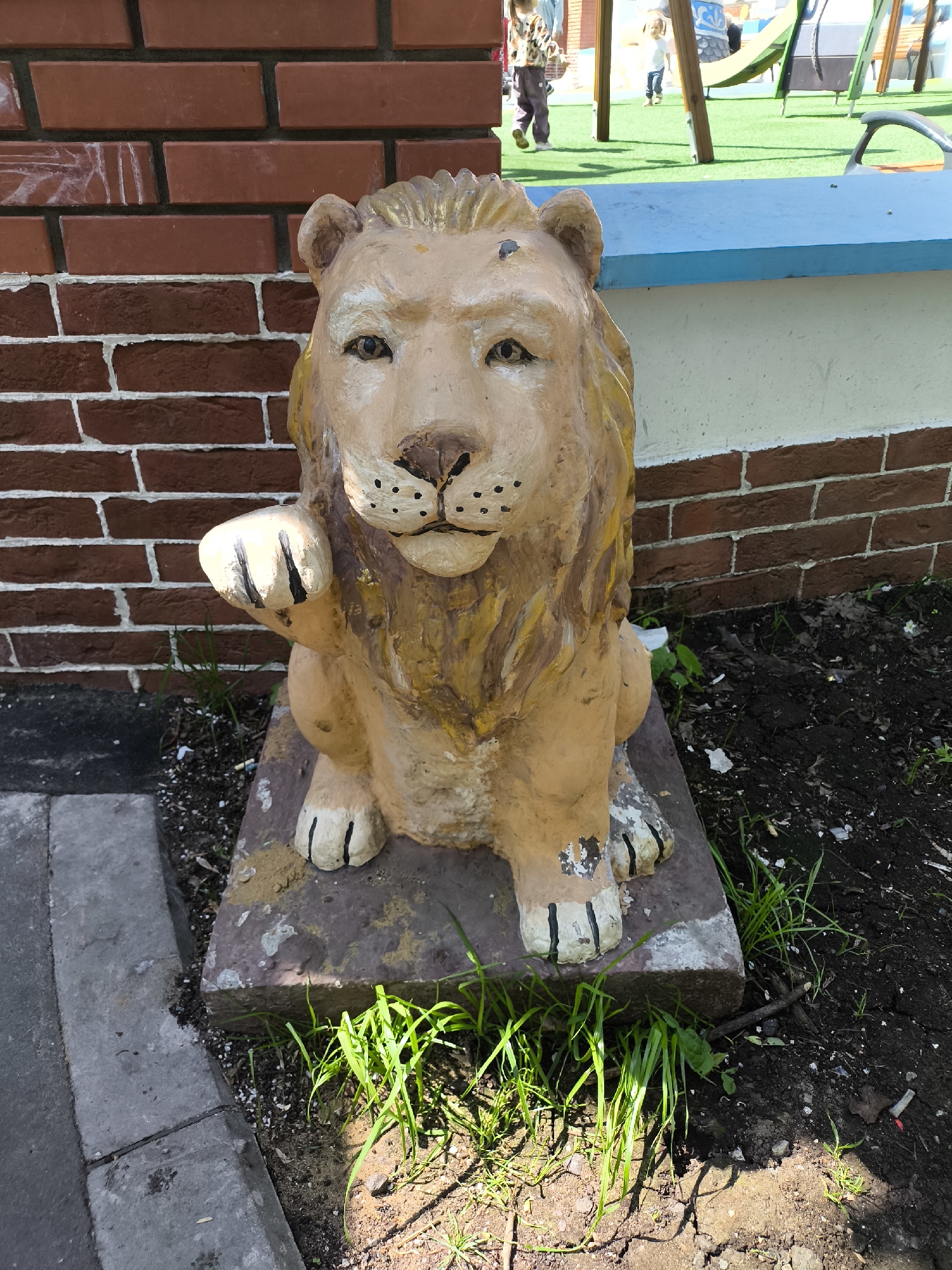 The image shows a tan, stylized lion statue sitting on a concrete base. It has a yellow-ish mane and black markings on its face and paws. The statue is located next to a red brick wall and a painted blue surface. In the background, there is a playground with children playing. Grass and soil surround the base of the statue. The location is Unknown, Unknown.
