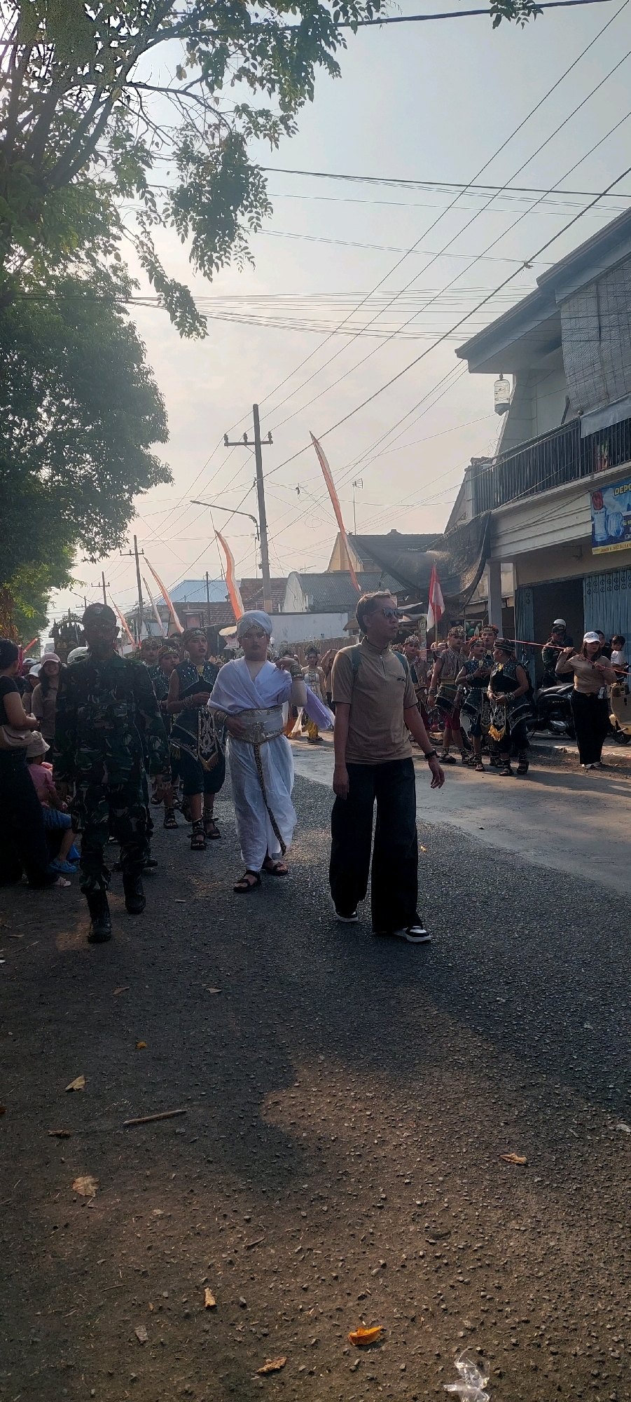 In Jogoyudan, Indonesia, a street procession is underway. A man in a camouflage uniform walks on the left, facing forward. To his right, a person in traditional white attire with a golden belt and headpiece is walking, also facing forward, and appears to be leading the procession. Following them are individuals in dark, ornate costumes, some carrying props. Spectators are visible on the sides of the street. The environment consists of a paved road lined with buildings and trees, with power lines overhead. The sky is bright and appears to be late afternoon or early morning, casting long shadows.