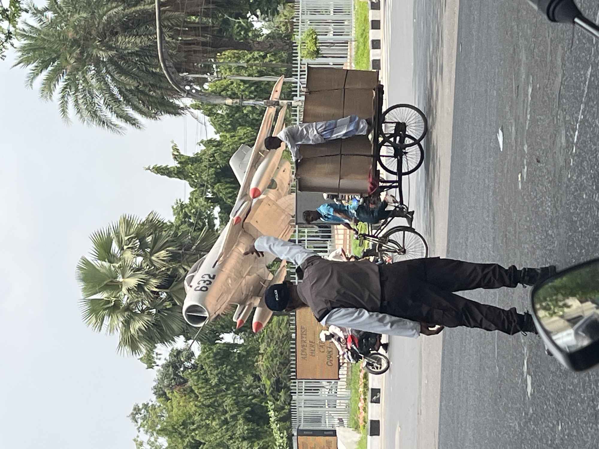 The image captures a scene in Dhaka, Bangladesh, featuring a MiG-21 fighter jet monument displayed outdoors. A man, possibly a security guard, stands in the foreground, pointing towards the monument. A three-wheeled bicycle loaded with boxes is positioned beside the monument, with two individuals tending to the goods. The environment includes trees, a metal fence, and a road.