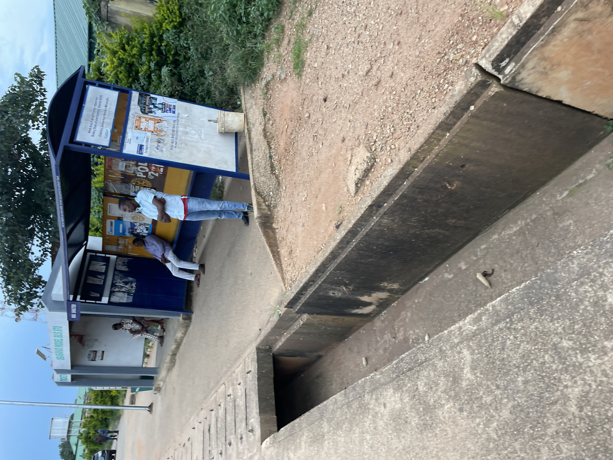 The image shows a bus shelter in Jos, Nigeria, labeled "Obasanjo Model Bus Sto". Two men are visible at the bus stop. One man in a light blue shirt and jeans stands with his arms crossed, looking to the left. Another man in a light colored long-sleeved shirt and white pants sits on the bench inside the shelter. There are several posters and signs displayed on the shelter, including one that reads "THIS BUS SHELTER WAS BUILT IN 2012/2013 YEAR BY: Rotary Club of Jos Central, District 9125". In the foreground, there is a concrete drainage channel running alongside the road. The ground is a mix of dirt and gravel, with some sparse vegetation. The sky is overcast.