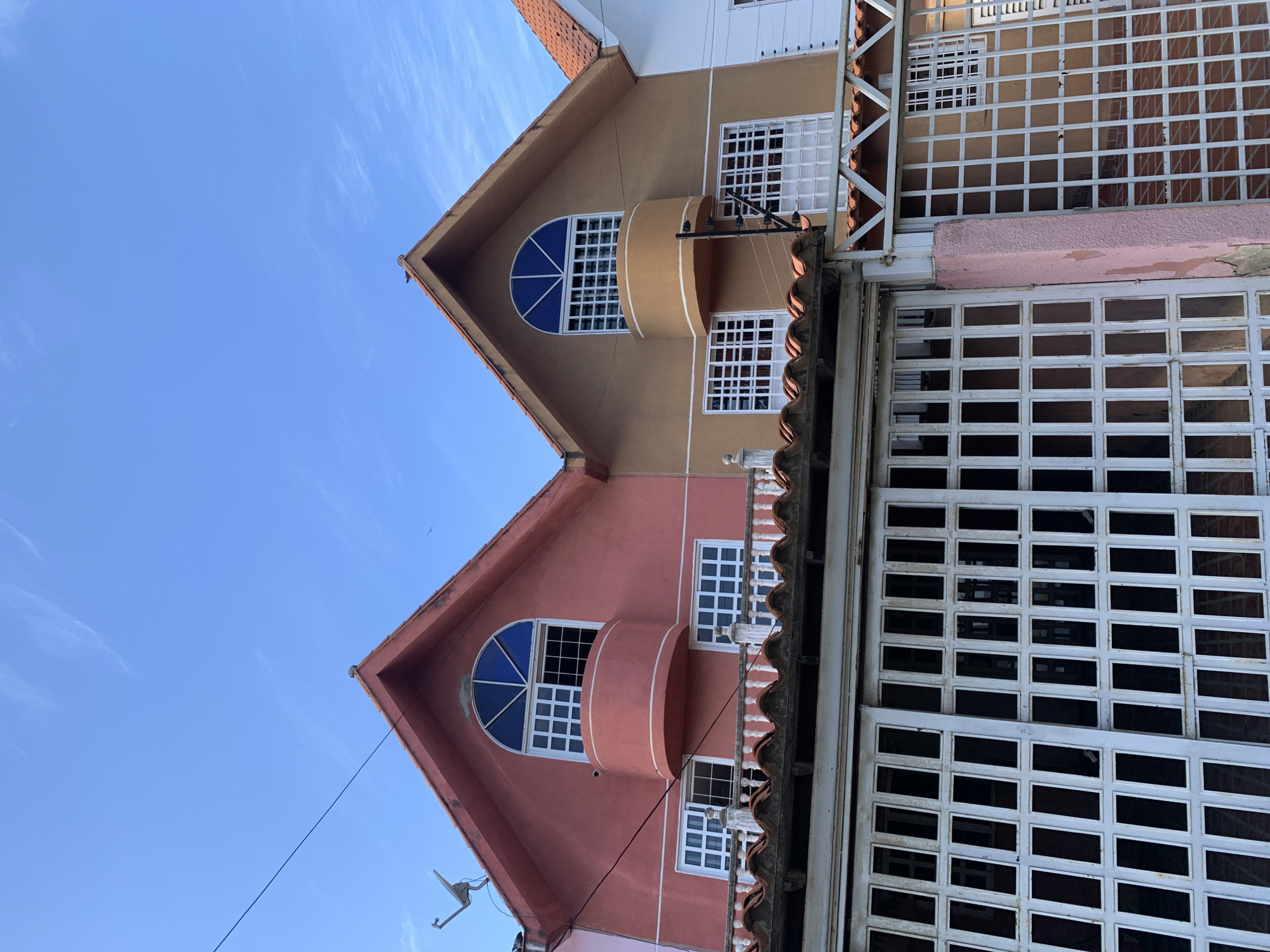 The image shows a section of a residential street in El Valle del Espíritu Santo, Venezuela. The focus is on the upper facades of two attached houses, one painted salmon pink and the other a light brown. Both houses have steeply pitched red-tiled roofs and feature similar architectural elements. Each has a distinctive arched window with blue glass at the top of the gable, below which are two rectangular windows with white grid-like security bars. A small, curved balcony juts out from the wall beneath the arched window of each house. A white balustrade runs across the front of the houses, just above street level. In the foreground, a white metal gate with a grid pattern obscures the lower portion of the buildings. The sky above is a clear, bright blue with a few wispy white clouds. A satellite dish is visible on the roof of the pink house, and a security camera is mounted on the brown house. The overall impression is one of a densely built residential area under a clear sky.