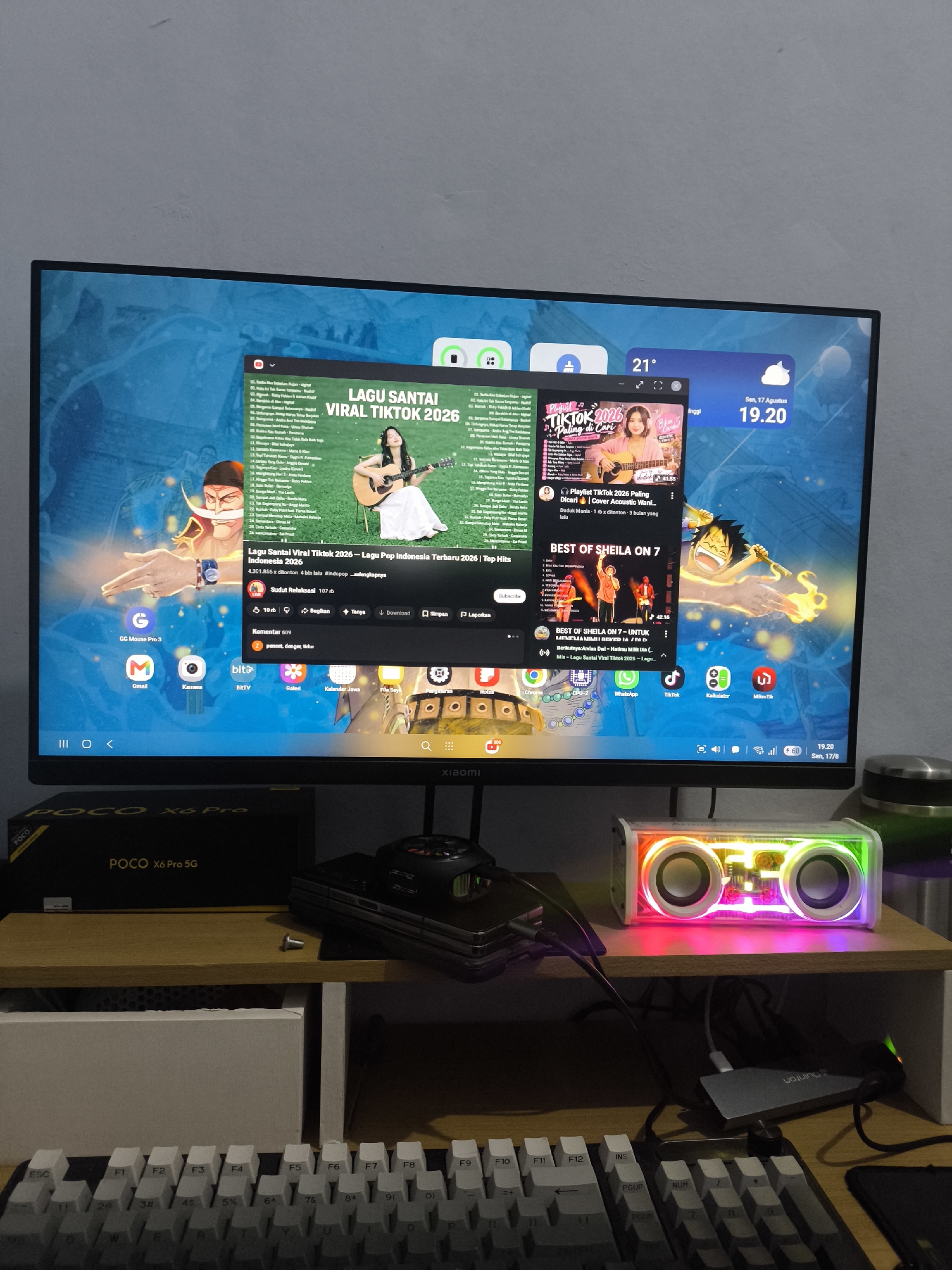 The image depicts a computer monitor displaying a YouTube video titled "LAGU SANTAI VIRAL TIKTOK 2026" and another video titled "BEST OF SHEILA ON 7". The video player interface, comments section, and related videos are visible. The monitor is positioned on a desk in what appears to be a bedroom or office setting.
On the desk, there is a keyboard, a portable speaker with RGB lighting, and a POCO X6 Pro 5G smartphone box. Cables are connected to the speaker and other electronic devices. The background shows a grey wall.
The image does not contain any information to place it in Kandangtepus, Indonesia.