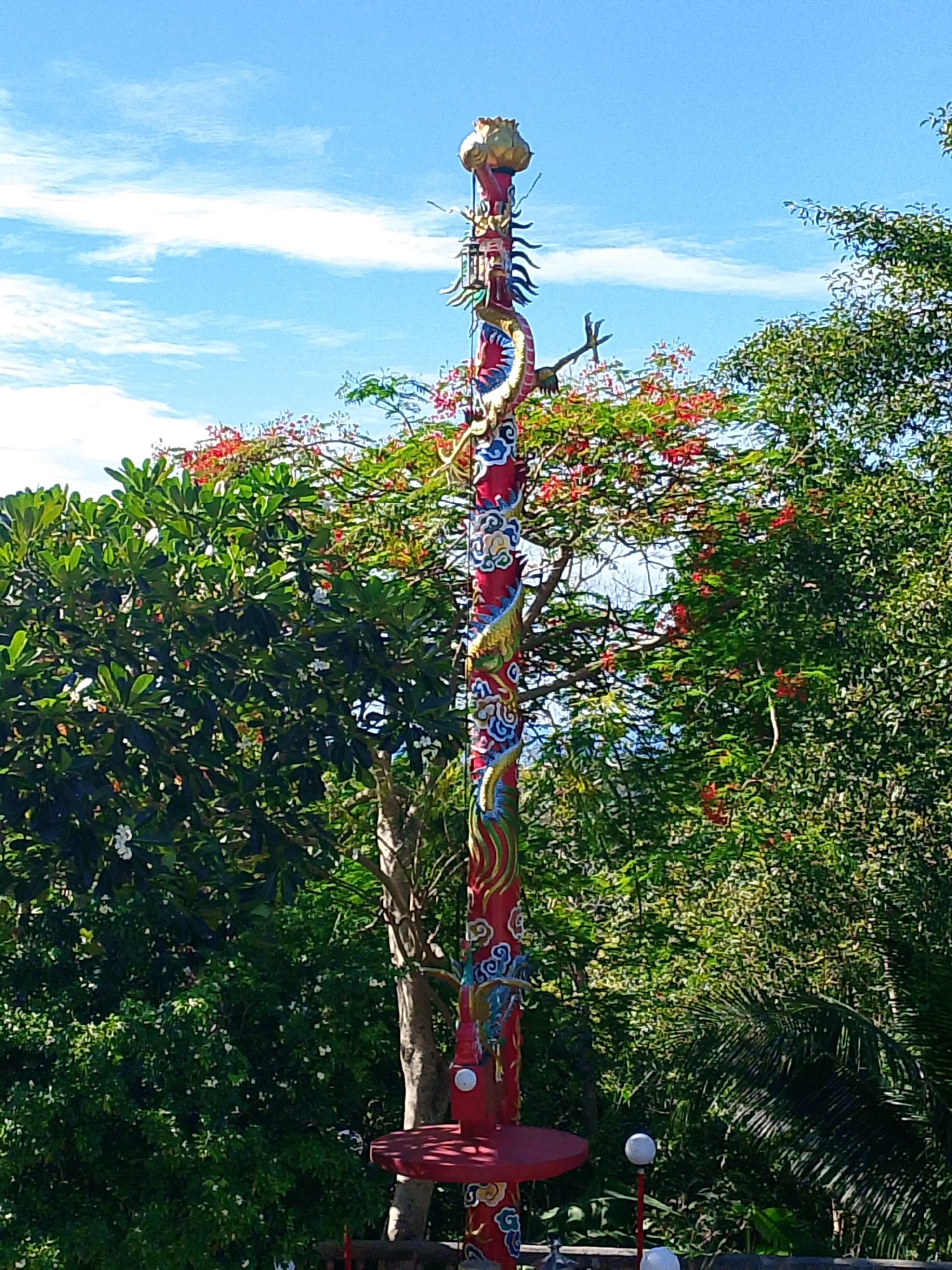 A tall, red, ornate pole with depictions of dragons and blue and white swirling patterns stands prominently in the foreground. The pole is topped with a golden finial. Surrounding the pole are lush green trees, some with vibrant red flowers. The sky is a clear blue with scattered white clouds. In the lower part of the image, there is a red platform attached to the pole, and two small white spherical lights on red poles are visible near the base. This scene appears to be an outdoor setting, possibly in a garden or park area in the city of Ko Pha Ngan, Thailand. There are no people or animals visible in the image.