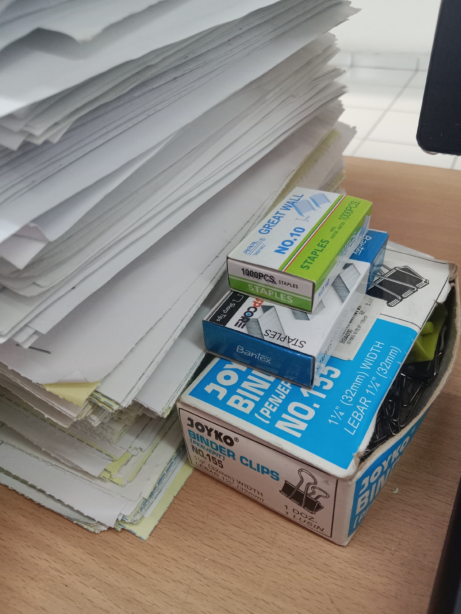 An eye-level, medium shot captures a stack of papers and office supplies on a wooden desk. The papers, mostly white with some yellow edges, are piled high on the left side of the frame, creating a backdrop for the main subject. In the foreground, three boxes of staples and binder clips are arranged in a slightly overlapping manner. The top box, green and white, reads "GREAT WALL NO.10 STAPLES 1000PCS". Beneath it, a blue and white box says "Banfex STAPLES". The bottom box, predominantly blue with white text, is labeled "JOYKO BINDER CLIPS NO.155 1 1/4\" (32mm) WIDTH LEBAR 1 1/4\" (32mm)". A black binder clip is visible within this box. The lighting is diffused, and the overall scene suggests an office environment. No people are present in the image.