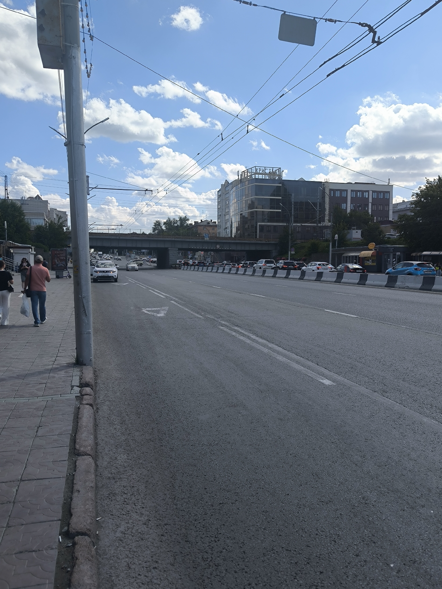 An outdoor, eye-level, vertical medium shot captures a street scene in Novosibirsk, Russia, under a partly cloudy sky. The foreground features a paved sidewalk with a decorative pattern, bordered by a concrete curb littered with cigarette butts. Adjacent to the sidewalk is a multi-lane asphalt road, where several vehicles are visible, including a white car and a blue car, indicating moderate traffic. In the middle ground, an overpass crosses the road, and beyond that, a modern, multi-story glass-fronted building with the inscription "КРИСТАЛЛ" (Kristall) is prominent, suggesting it is a business or commercial establishment. The background includes more buildings and trees, with overhead wires and poles suggesting an urban electrical or transport system. Two individuals are walking on the sidewalk in the mid-left of the frame, one carrying a white bag. The overall impression is an everyday urban environment.