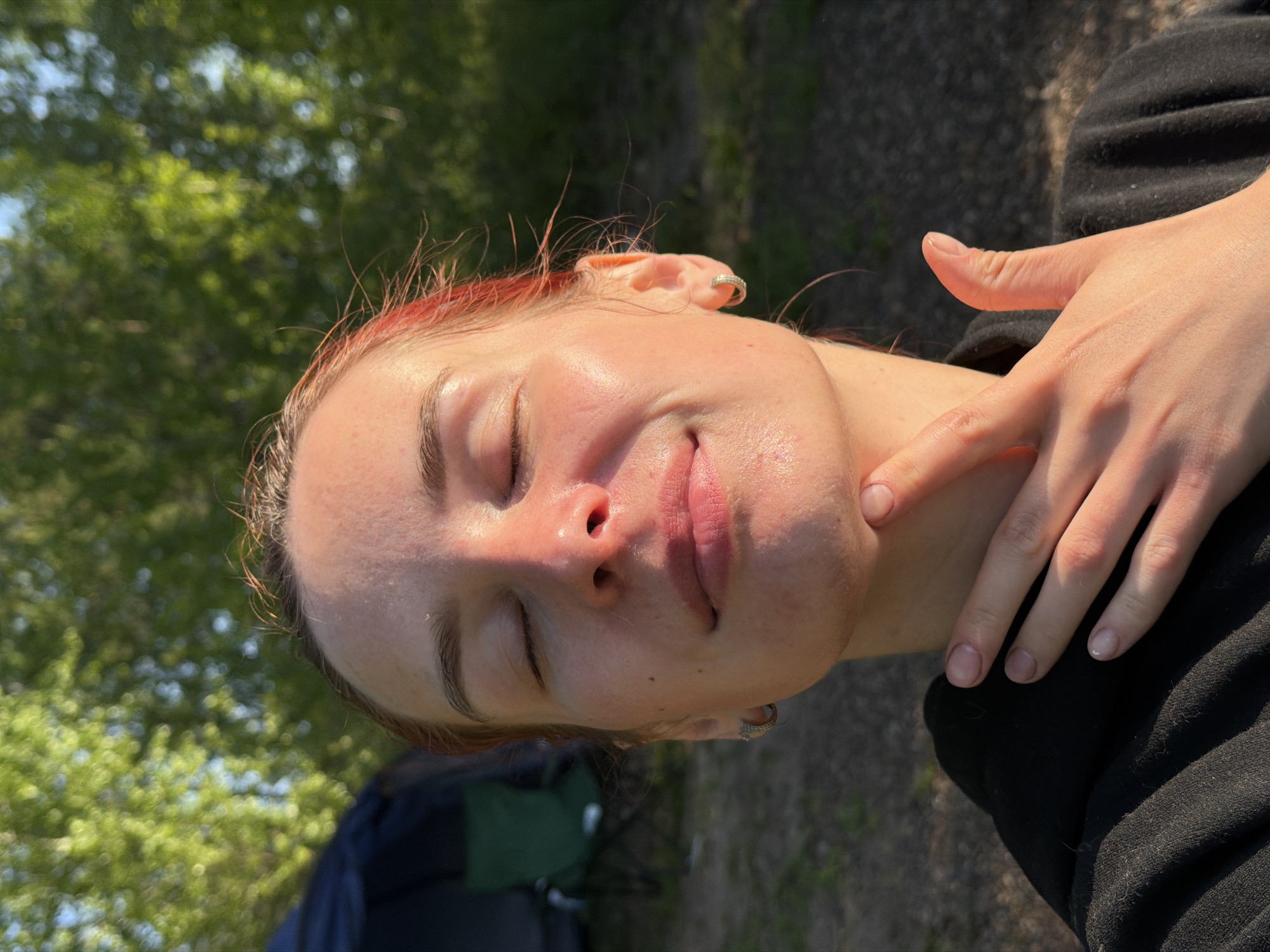 A close-up shot of a woman, seemingly outdoors, with her eyes closed, is presented. She is fair-skinned with a hint of pink in her hair. She is wearing a black top. Her right hand is near her face, with her index finger pointing towards her chin. The background shows blurred greenery and possibly a tent. The exact location is not identifiable as Noviy, Russia, from the visual data.