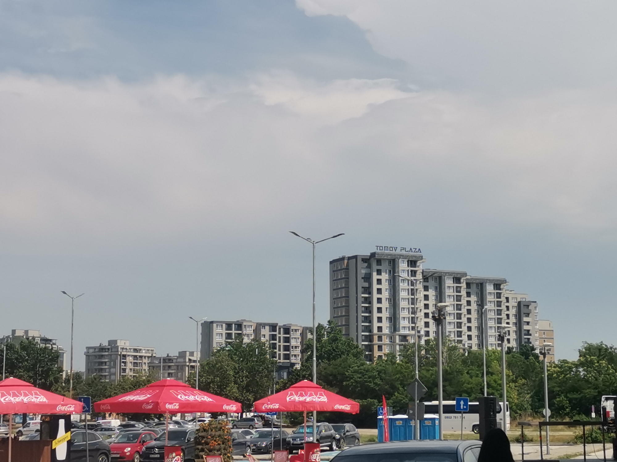 This image captures a wide outdoor urban scene in Plovdiv, Bulgaria, specifically featuring the Tomov Plaza complex, under a bright, overcast sky. In the immediate foreground, three large, red circular umbrellas, each branded with "Coca-Cola" in white lettering, are prominently displayed. Beneath the left-central umbrella, a yellow sign with "Schweppes" is visible. A paved parking lot extends across the mid-ground, populated by numerous parked vehicles of various makes and models, predominantly dark-colored sedans, with one distinct red car in the center-right. Modern, silver-colored streetlights with double luminaires are spaced throughout the area.
Towards the right side of the image, a row of three blue portable toilets is visible. Behind these, a white bus, marked with the phone number "0883 781 714" and a blue upward-pointing arrow sign, is partially obscured. Dense green trees and bushes act as a natural separation between the parking area and the background structures. In the background, a series of large, multi-story buildings define the skyline. The most prominent complex, centrally located, is identified by "TOMOV PLAZA" spelled out in white letters on its roofline. This complex comprises several interconnected buildings with a uniform modern architectural style, featuring gray and white facades and numerous rows of rectangular windows. Additional similar residential or commercial buildings are visible to the left and right of the Tomov Plaza complex, receding into the distance. A person with dark hair is partially visible in the bottom right corner of the frame. The sky above is a consistent light gray, indicating full cloud coverage with no direct sunlight.