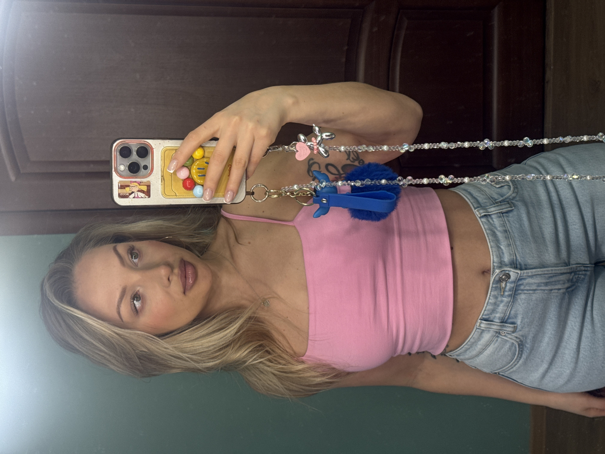 A woman is taking a selfie in front of a dark brown paneled door and a green wall. She is holding a smartphone with a decorative case and beaded strap. The case features cartoon characters, colorful beads, and a yellow smiley face. The woman is wearing a pink tank top and light blue jeans. Her hair is blonde and styled with highlights. She has a tattoo on her upper arm. The location is not specified, and there is no information in the image to indicate it is Moscow, Russia.
