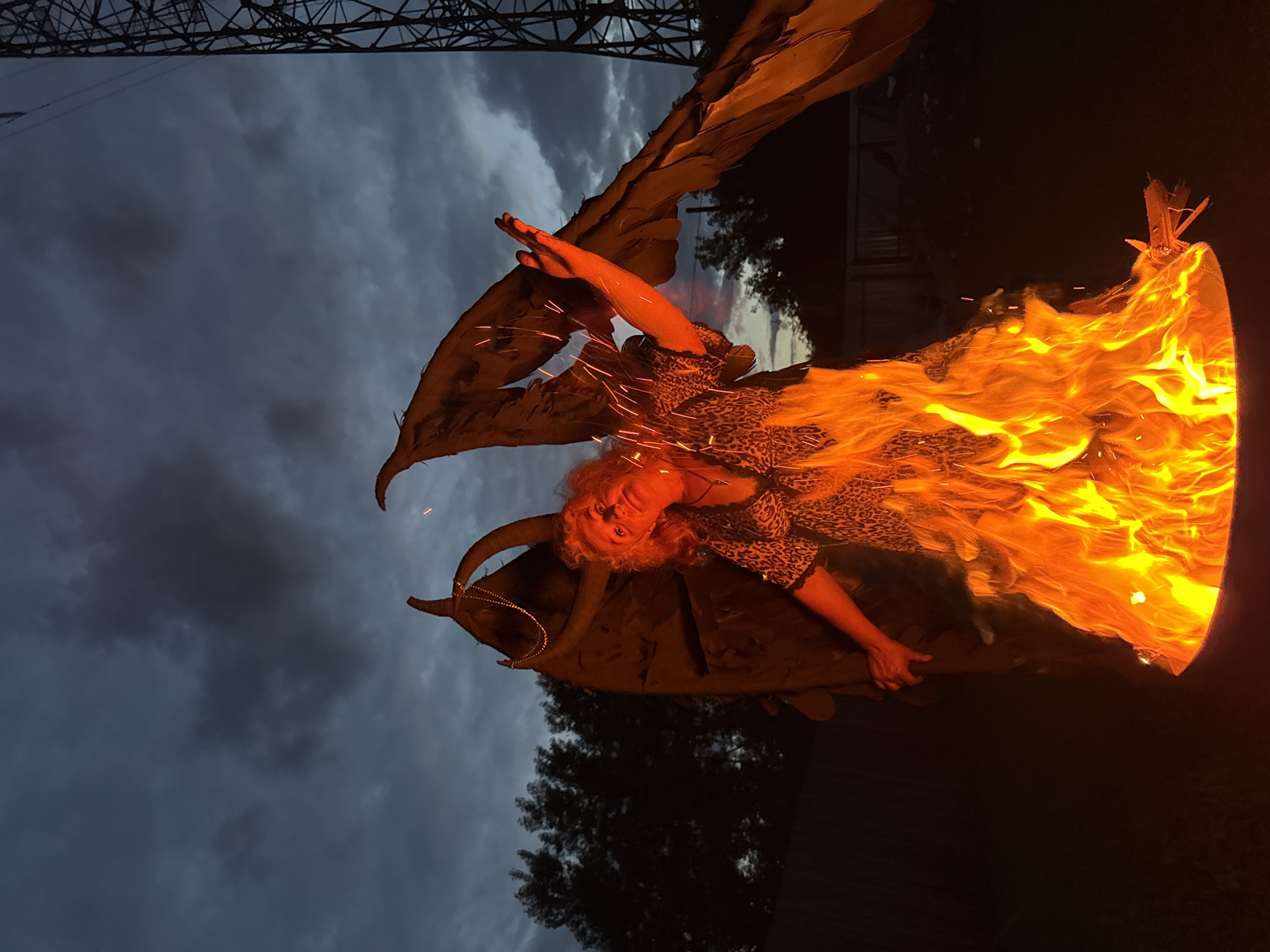 A person with their face illuminated by red light stands within a large bonfire. They have curly red hair and are wearing a leopard-print dress. To their sides are large, dark wings, one of which is angled upwards with a hand outstretched. Sparks are visible in the air around the person. The bonfire is contained within a dark metal drum. In the background, silhouetted trees and a tall metal structure, possibly a pylon, are visible against a cloudy, dark sky. The scene appears to be outdoors, likely in a backyard or open area in the city of Chaikovsky, Russia.