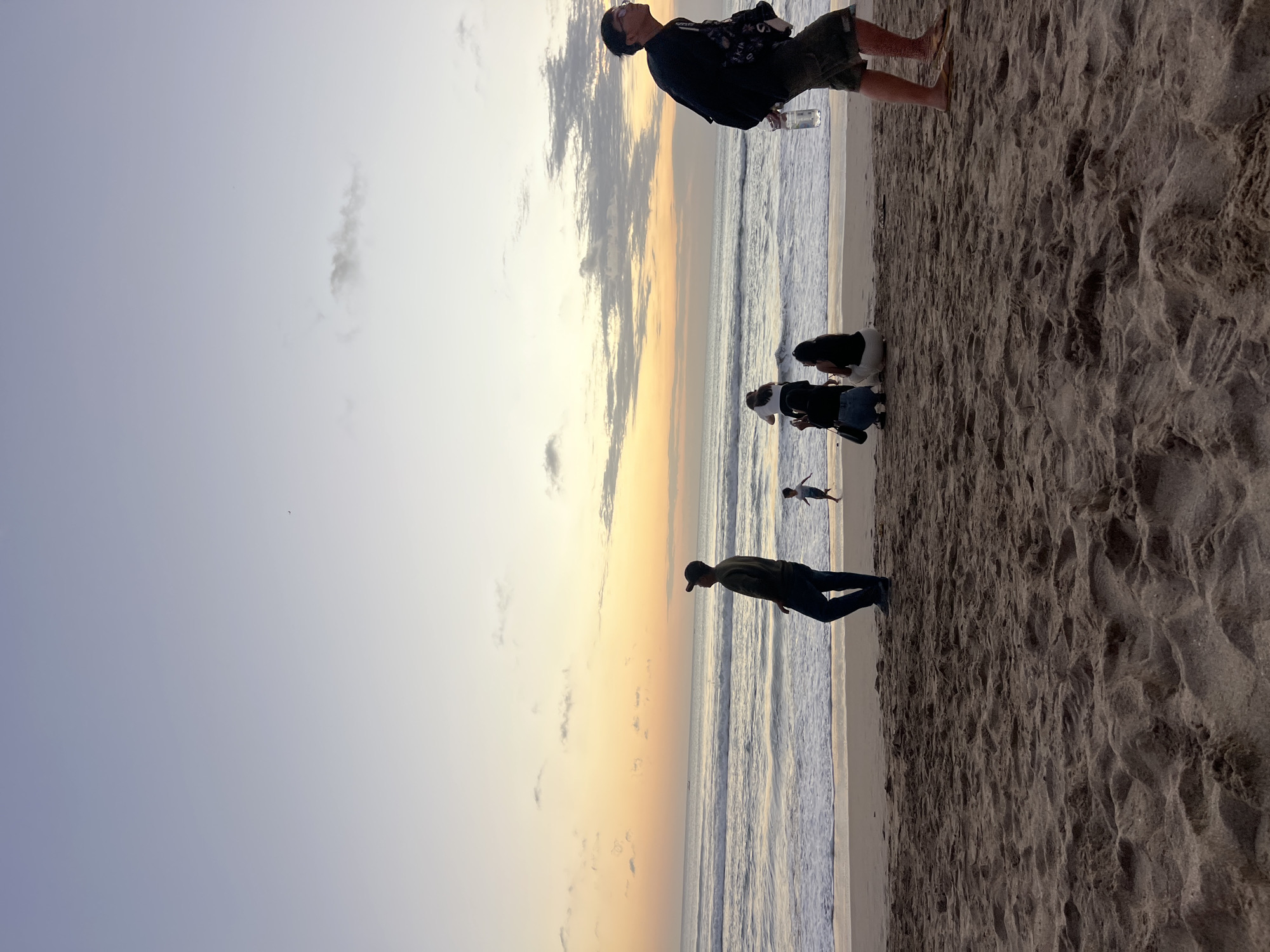 The image depicts a serene beach scene in Kuta, Indonesia, during what appears to be either sunrise or sunset. The sky transitions from a soft lavender to a warm gradient of orange and yellow as it meets the calm ocean. Gentle waves roll onto the sandy shore, which is covered in the fine grains of sand typical of Kuta's beaches.
Several individuals are present, scattered across the shoreline. In the foreground, a man in dark clothing and a cap stands facing the ocean, his gaze directed towards the horizon. Further back, a small child runs along the water's edge. To the right, another man walks away from the camera, carrying a water bottle and a backpack, wearing shorts and flip-flops. In the mid-ground, a group of three people, two women and one person who appears to be another woman, are seated on the sand. They are facing the ocean, with one person appearing to take a photograph with a phone. The overall atmosphere is peaceful and contemplative, with the natural beauty of the beach setting dominating the scene.