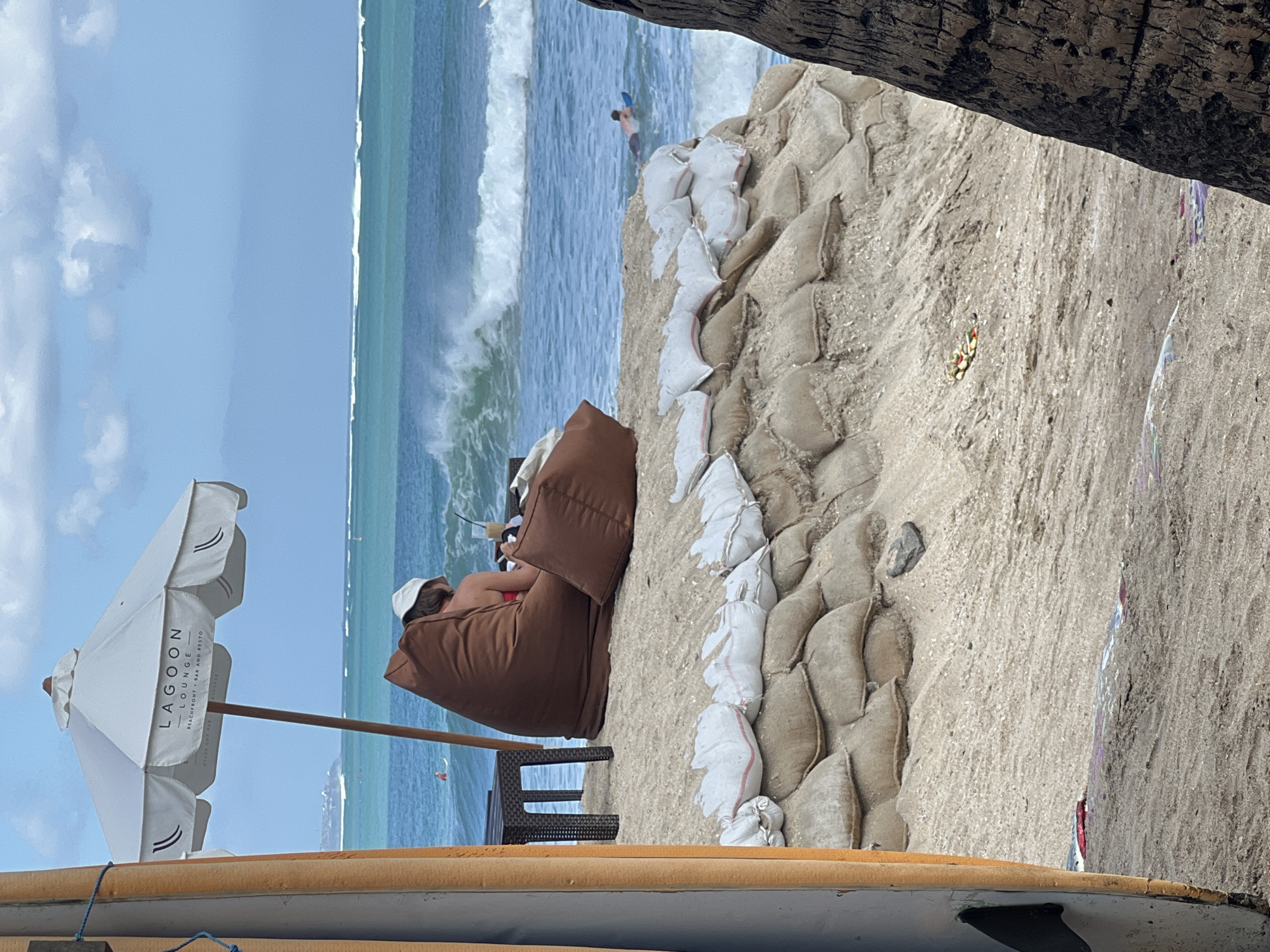 An overcast day is depicted on Kuta Beach, Bali, Indonesia. The image shows a sandy beach with a line of sandbags reinforcing the shoreline. In the mid-ground, a person sits in a large, brown beanbag chair under a white umbrella that reads "Lagoon Lounge". The person is wearing a white hat and appears to be looking at a phone or book. A small table with a dark surface is next to the beanbag chair. The ocean is visible in the background with moderate waves. A lone surfer is in the water. Part of a surfboard is visible on the left side of the frame, and a palm tree trunk is on the right.