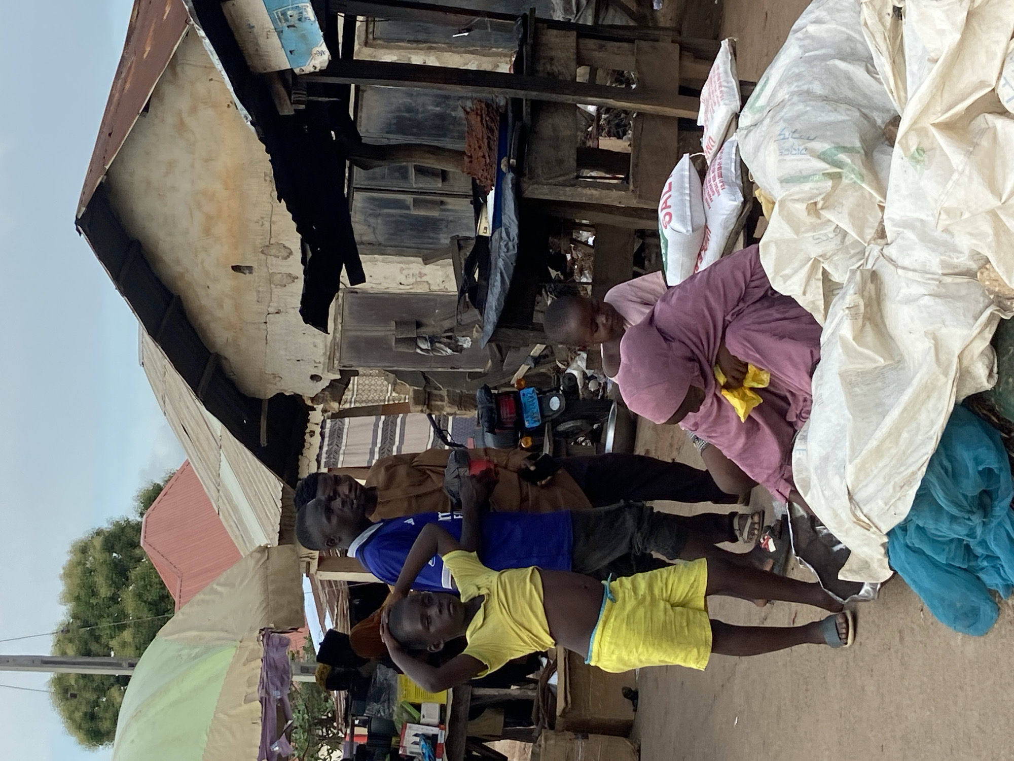 An outdoor, medium shot, eye-level, horizontal photograph taken in Mararaba, Nigeria, shows six people in an open-air market during the day under an overcast sky. The image captures a bustling street scene with various elements of daily life.
In the foreground, a young child wearing a yellow t-shirt and shorts stands facing the camera, with hands on their head. To their left, a man in a blue t-shirt and dark shorts stands behind them, looking towards the right side of the frame. Behind him, another man in a brown long-sleeved shirt and dark pants is visible, also looking in the same direction. Further to the right, a person in a pink or purple garment is seen hunched over, seemingly attending to something, with yellow fabric in their hand. Two other individuals are partially visible: one person wearing dark shorts stands next to the person in pink, and another is seated near the bags on the far right.
The background features a mix of structures and market goods. On the right, large white and beige sacks, possibly containing grains or other market items, are stacked. Some of these bags have visible text, such as "SAL" and "UN". A blue piece of fabric is also seen near the bags. In the mid-ground, a black motorcycle is parked. The buildings in the background appear to be constructed with corrugated metal roofs and weathered walls, suggesting a functional, perhaps aged, urban environment. There are also visible wooden beams and various items hanging, such as black clothing and other fabrics, indicative of a market stall or residential area. On the far left, a large, light-colored umbrella is partially visible, and a lush green tree can be seen in the distance against the light sky. The ground is a dirt or unpaved surface. The overall atmosphere appears busy but calm.