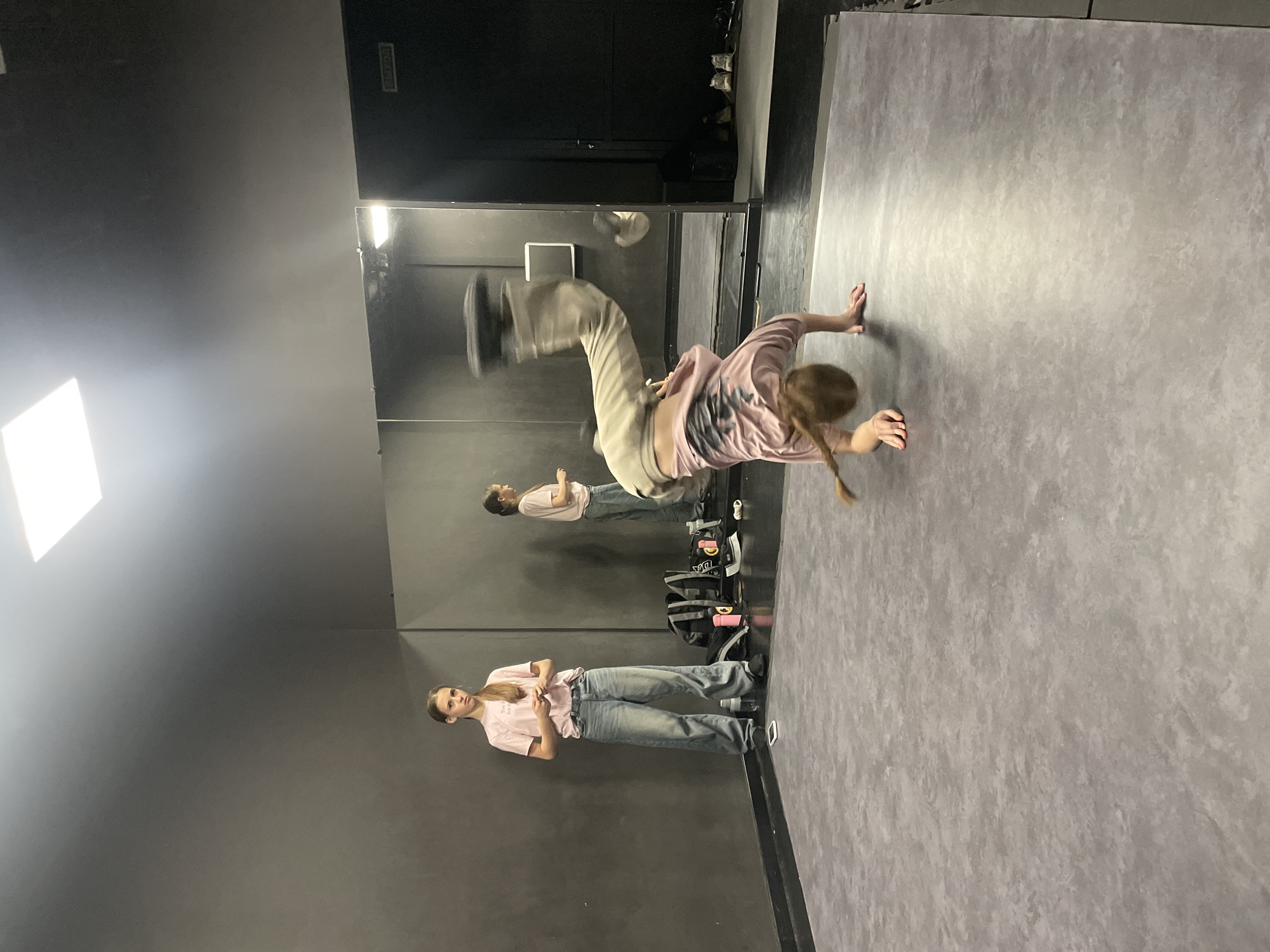The image shows a young woman in the midst of a breakdancing move, possibly a handstand or related acrobatic pose, in a dance studio. She is wearing a pink shirt and baggy pants, and her hair is in a braid. A mirror reflects her image and another person standing casually, dressed in a similar outfit. The studio has dark walls and a textured gray floor. Various items like bags and water bottles are visible near the mirror. There is no way to identify the precise location within the city of Krasnoyarsk, Russia without additional information.