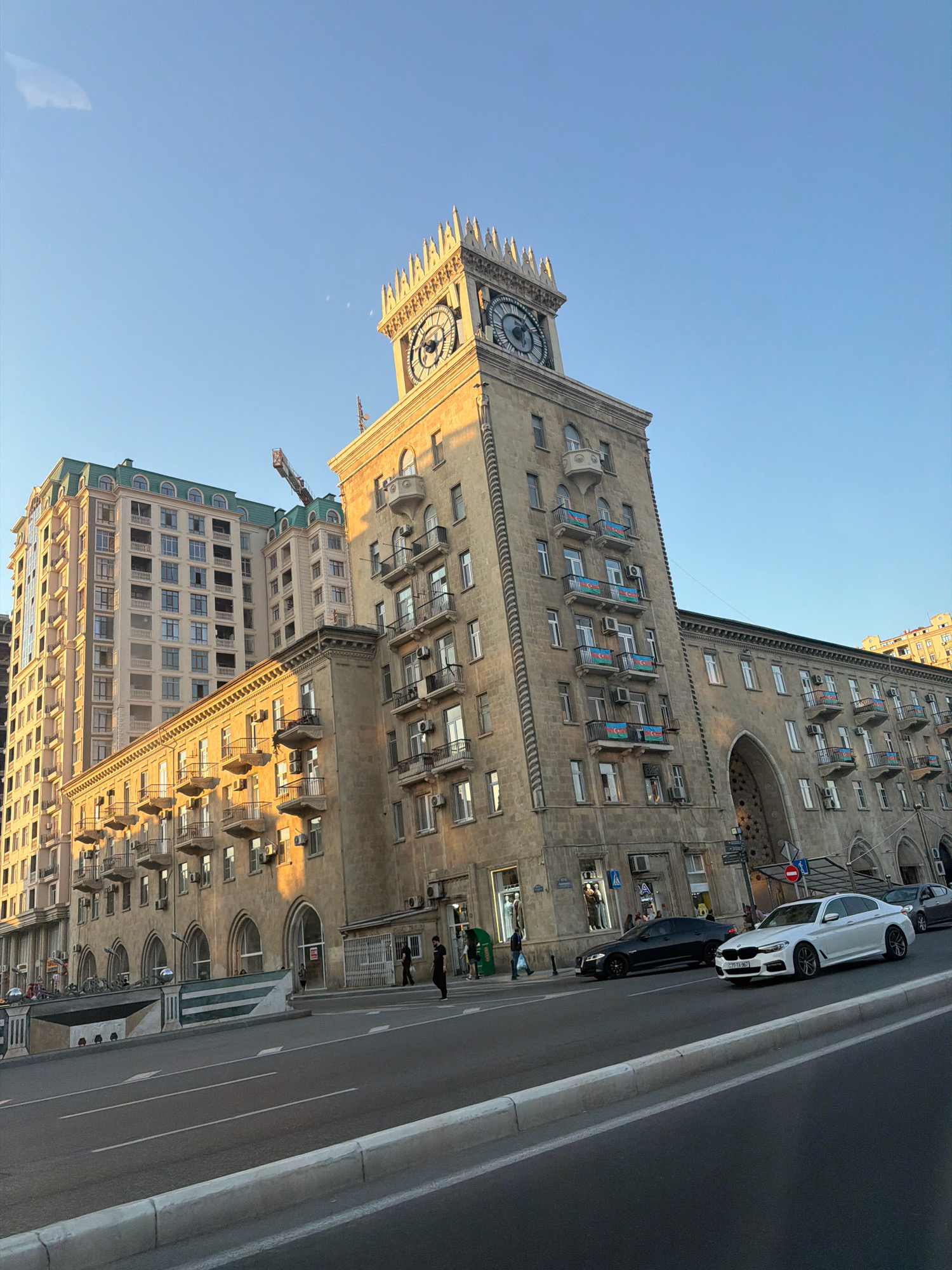 The image captures a street scene in Baku, Azerbaijan, featuring the iconic Clock Tower building and adjacent apartment complexes under a clear blue sky. The sun is casting a warm, golden light, highlighting the architecture. In the foreground, a multi-lane road shows traffic with a white BMW sedan and a black Jaguar driving by. Pedestrians can be seen on the sidewalk near shop entrances. The Clock Tower, a prominent landmark, displays its large clock face and decorative crenellations. Several balconies on the buildings are adorned with Azerbaijani flags. Construction cranes are visible in the background, suggesting ongoing development or renovations. The scene depicts a vibrant urban environment in Baku.