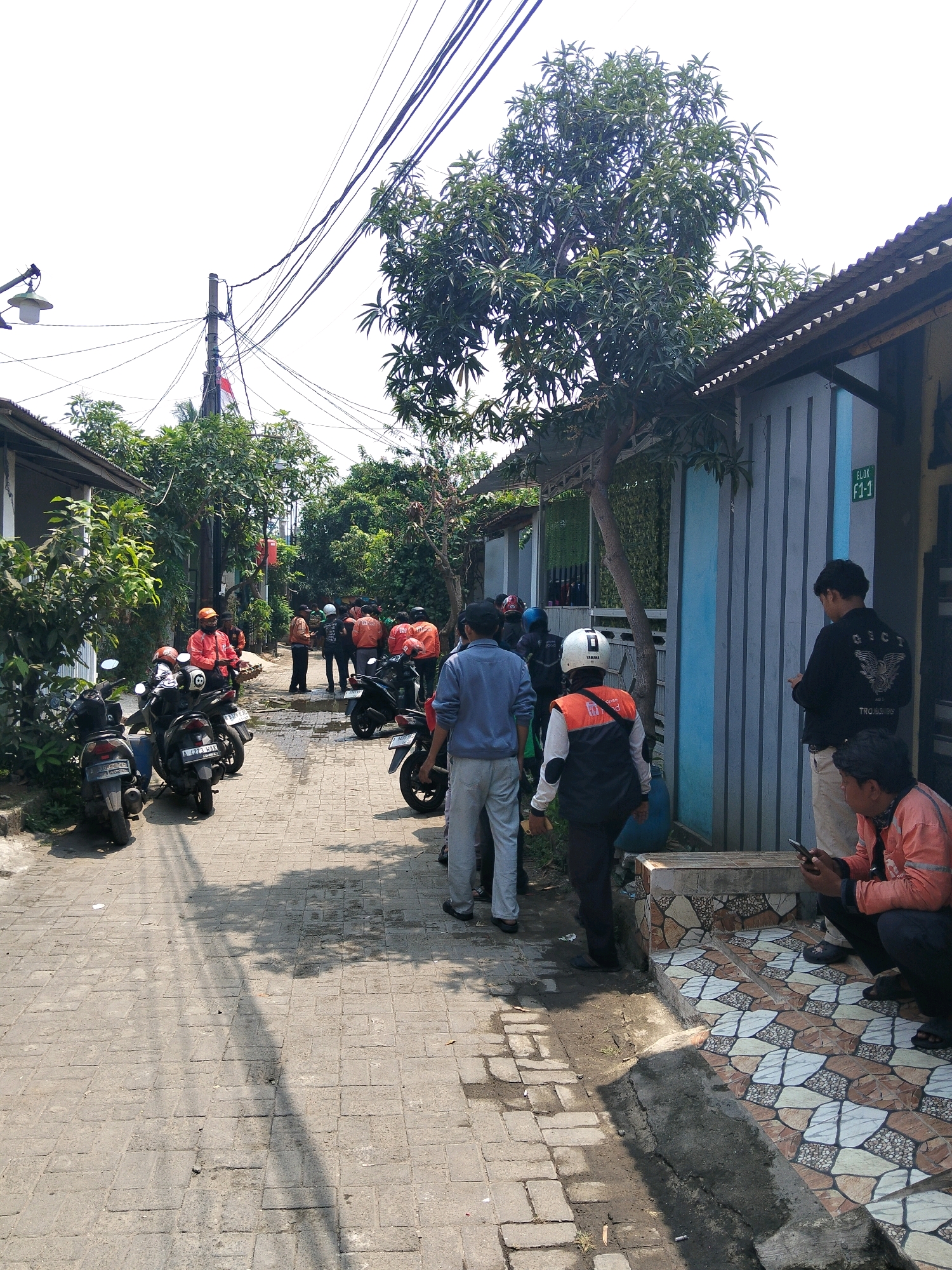 This is a wide shot of an alleyway in Rajegmulya, Indonesia. The alleyway is paved with bricks and lined with houses and trees. Several people are gathered in the alleyway, many of whom appear to be wearing orange jackets and helmets, suggesting they may be delivery drivers or workers. They are standing around, talking, and looking at their phones. Some motorcycles are parked in the alleyway. The sky is overcast, and the sun is casting shadows on the ground. The scene appears to be during daylight hours.