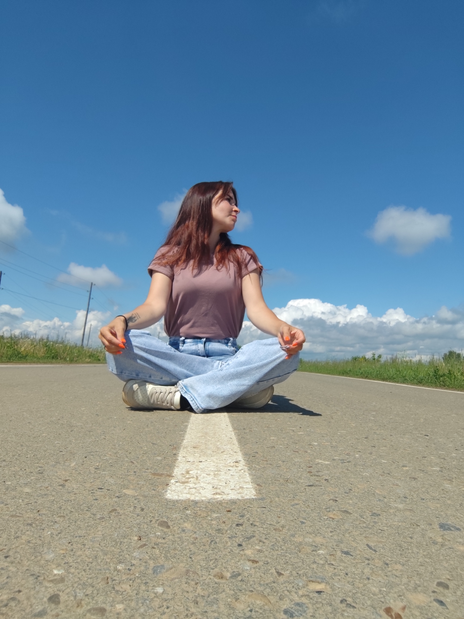 A young woman with long, reddish-brown hair is depicted sitting cross-legged on an asphalt road under a bright blue sky. She is wearing a light mauve t-shirt, light blue loose-fitting jeans, and white sneakers. Her hands rest on her knees, palms facing upwards, with her fingernails painted a vibrant orange. A dark bracelet or band is visible on her left wrist, along with a small tattoo on her left forearm.
The woman is positioned on a distinct white painted line running down the center of the road, and she gazes calmly towards her right with a serene and thoughtful expression. The shot is taken from a low angle, emphasizing the expanse of the sky, which is dotted with scattered white, fluffy clouds.
The setting appears to be a rural or semi-rural area in Фоки, Russia. Green grass and fields line both sides of the road, extending into the distance. Faint utility poles or power lines can be seen in the far left background. The strong, clear light indicates a sunny day, likely midday or early afternoon, casting soft shadows on the road surface. There is no visible text in the image.