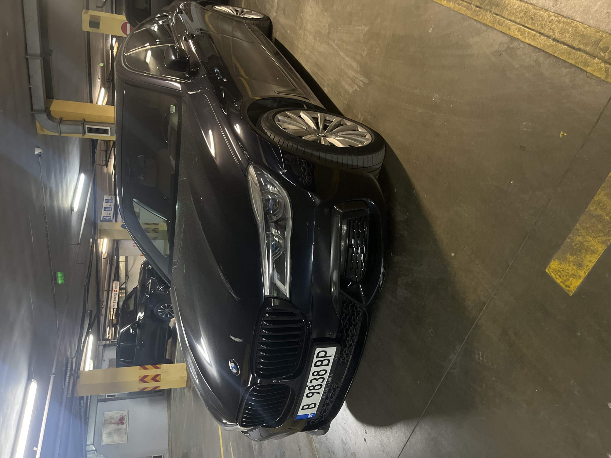 The image shows a dark BMW sedan parked at an angle in an indoor parking garage in Varna, Bulgaria. The car's front end is prominently displayed, including the grill and license plate, which reads "B 9838 BP" with "MCAR VARNA" on the bottom. The garage has painted yellow lines and support pillars. Other cars can be seen parked further in the background. The lighting is provided by overhead fluorescent lights.