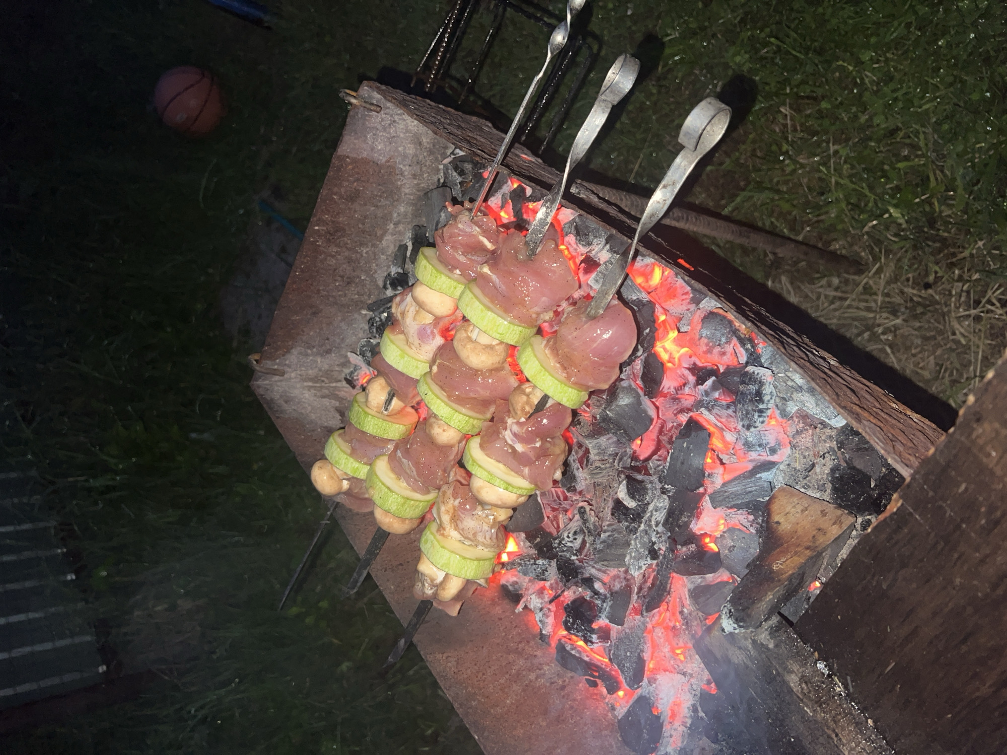 The scene depicts an outdoor nighttime grilling activity in the city of Chaikovsky, Russia. A rectangular, dark metal brazier, appearing weathered or rusted, contains a bed of glowing red and orange hot coals mixed with black charcoal. Five metal skewers are placed horizontally across the top edges of the brazier, positioning their contents directly above the embers. Each skewer holds alternating pieces of raw, light-pink meat, circular slices of green zucchini, and whole light-brown mushrooms. Light smoke is observed rising from the coals. The brazier rests on a dark green grassy surface, which exhibits a subtle sheen suggesting dampness or dew. In the upper left background, a dark, vertically textured fence or wall is partially visible. An orange basketball, appearing deflated or partially deflated, rests on the grass in the upper right portion of the frame. No individuals are present in the image.