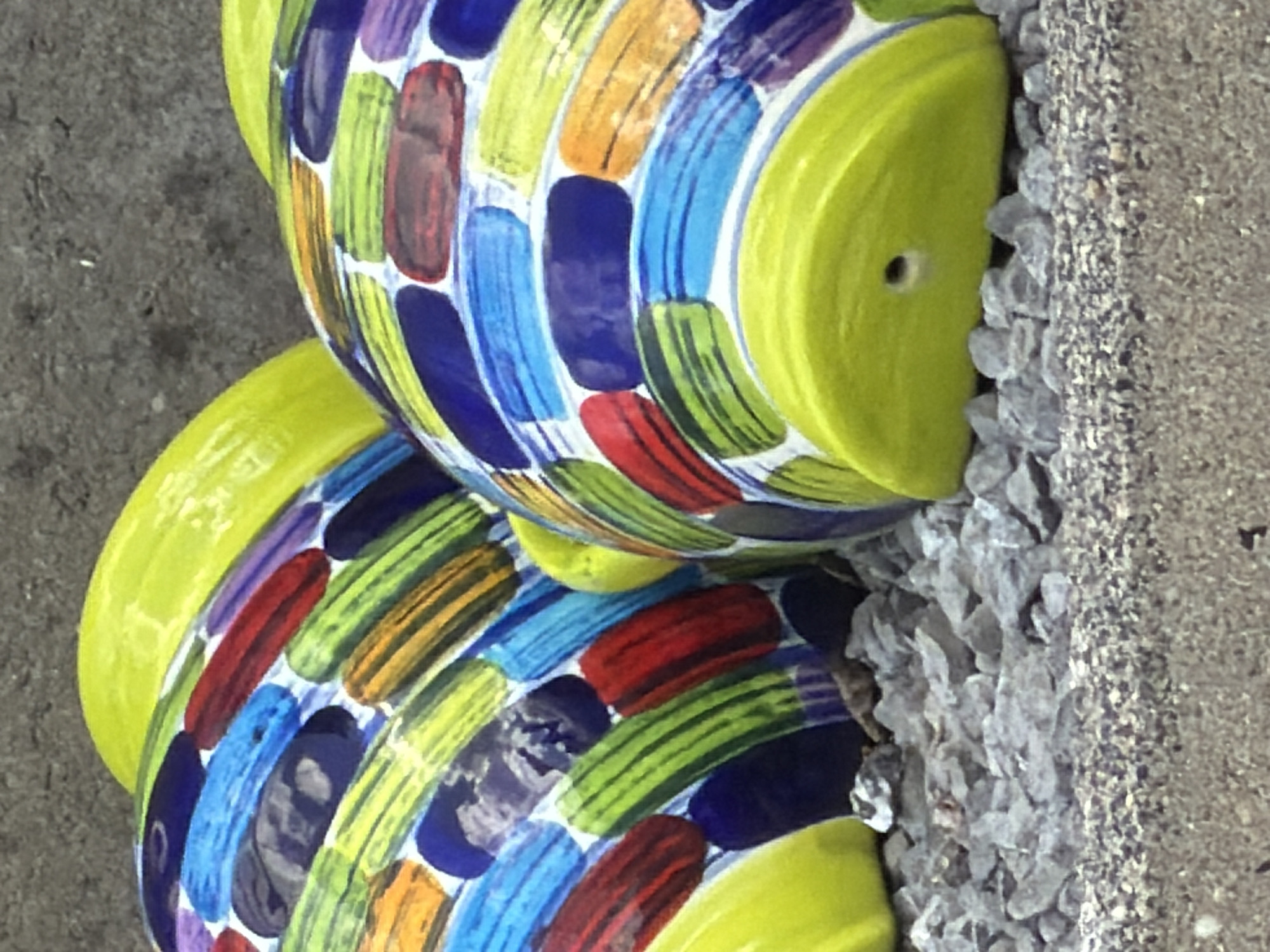 Two ceramic fish planters, painted with colorful stripes of red, blue, green, orange, and purple, are stacked vertically. They have yellow "heads" and are positioned next to a gravel and concrete surface. The location is Esch-sur-Alzette, Luxembourg.