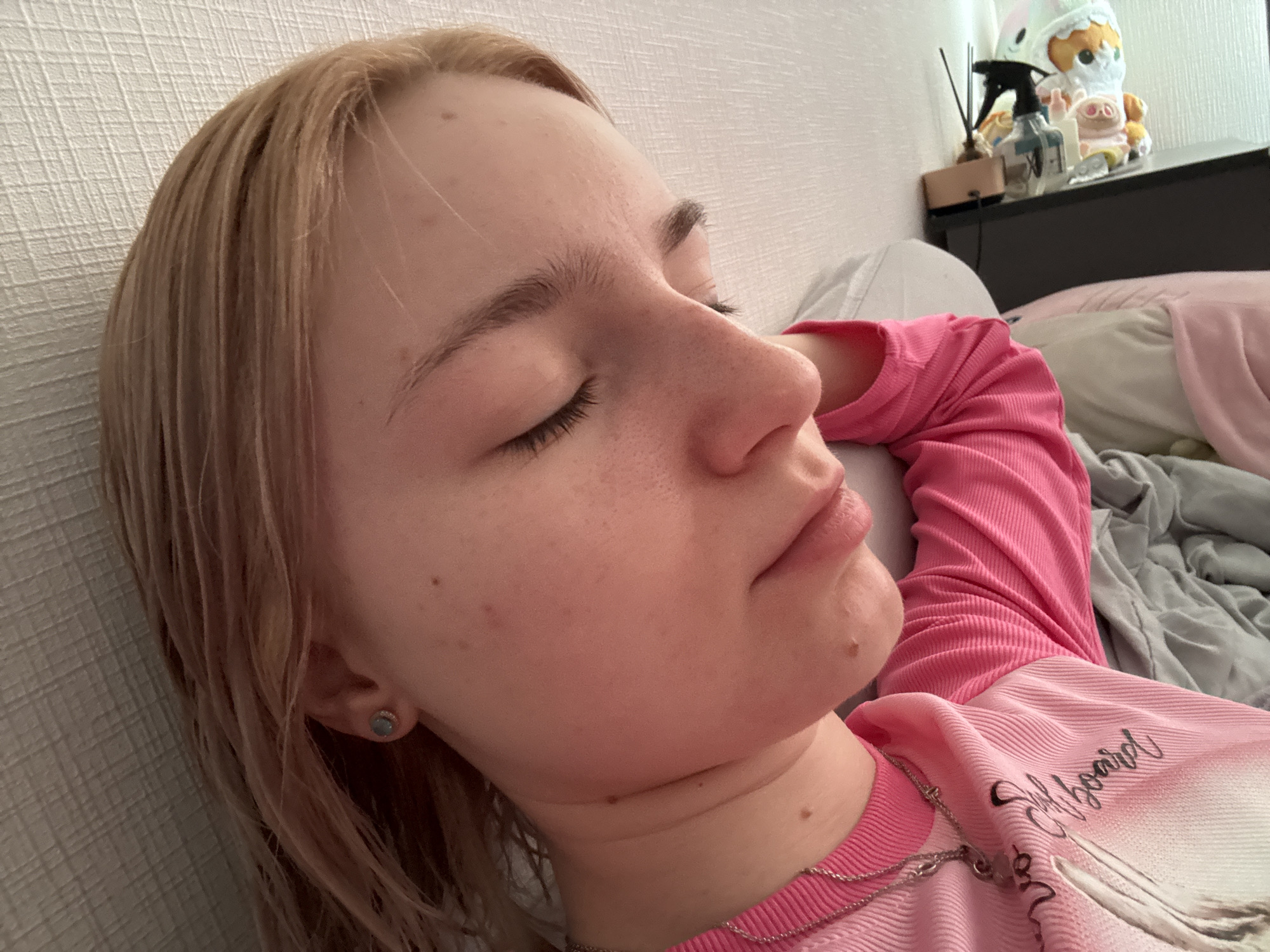 The image depicts a young individual, appearing to be female, with light blonde hair, resting or sleeping on their right side with eyes closed. The scene is located indoors, presumably a bedroom, in the city of Unknown, Russia. The individual has fair skin with multiple small moles on the forehead, cheek, and neck. A small, light blue or grey stud earring is visible in the right earlobe. They are wearing a long-sleeved, ribbed pink top. The top features a partial white graphic and text "Srfboard" on the chest area. A silver chain necklace with a pendant rests on the individual's collarbone. The person is lying on white and grey bedding, against a textured white wall. To the right, a dark nightstand is visible, holding several objects: a black spray bottle, a decorative diffuser with dark reeds, a small plush pig toy, and a larger, multi-colored plush toy resembling a creature with a hat.