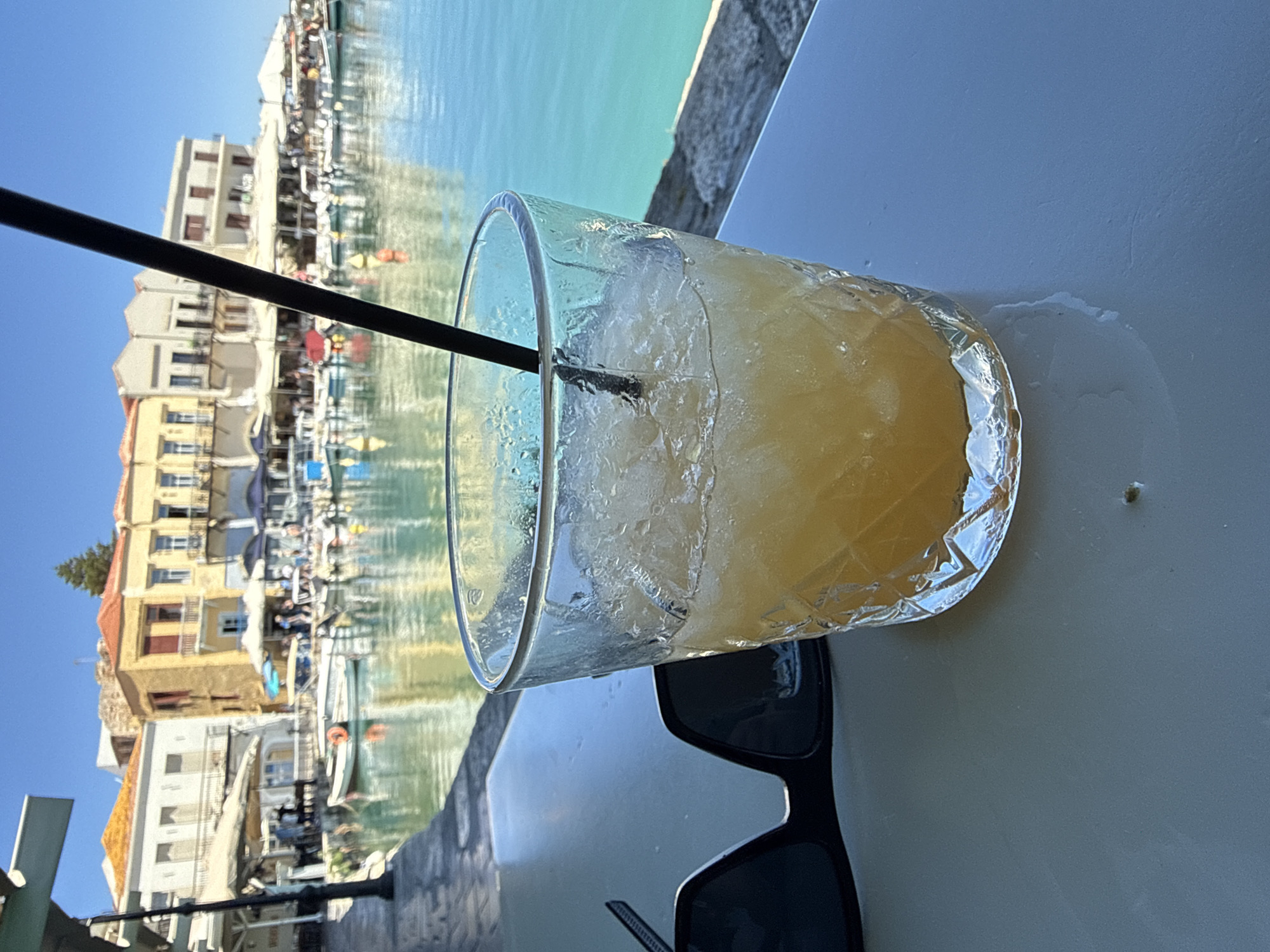 The image depicts a close-up view of a glass containing a yellow beverage with ice and a black straw, placed on a table. To the left of the glass are a pair of black sunglasses. The background reveals a harbor scene in the city of Rethymno, Greece. Several boats are docked in the turquoise water, and a row of buildings with traditional architecture lines the waterfront. The sky is clear and blue. The scene appears to be a snapshot of a relaxed moment at a waterfront cafe or restaurant.
